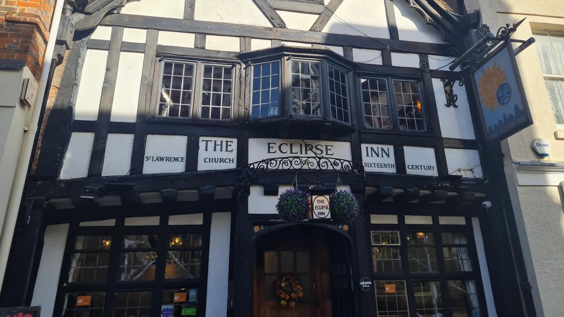 A half-timbered three-storey building is sandwiched between two lighter coloured buildings. The first floor has a large bay leaded window and beneath it reads the words 'The Eclipse Inn'.