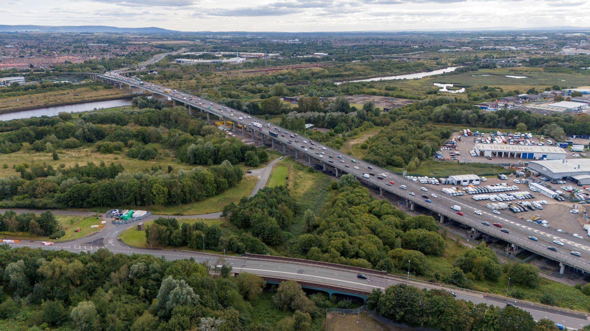 A six-lane flyover runs from bottom right to top left through a wooded landscape and across a river.