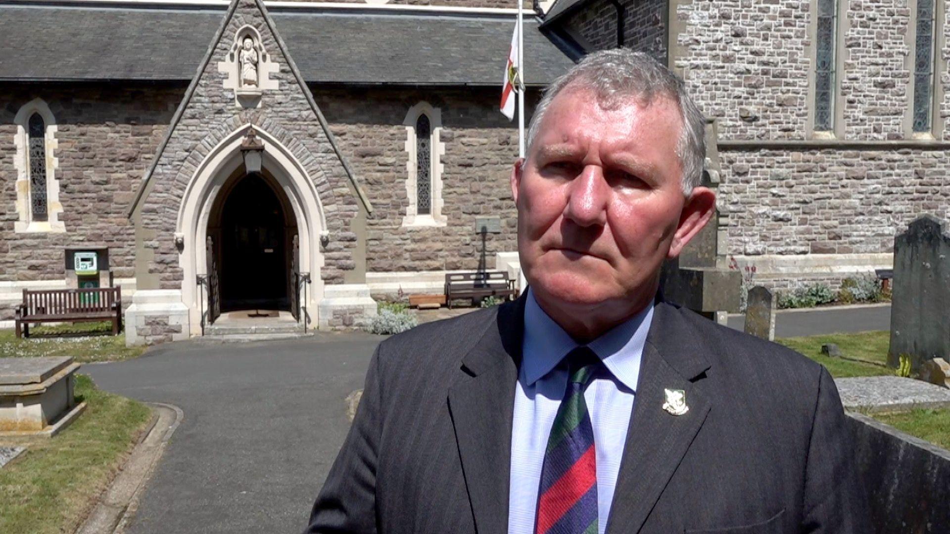 A man in a suit stood in a church graveyard. There is a flag in the background.