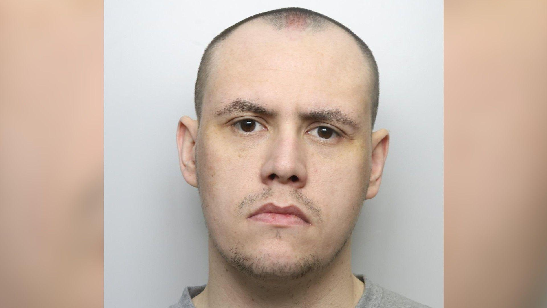 A police custody picture of Adam Martin. He has a shaved head and stubble and is wearing a grey jumper