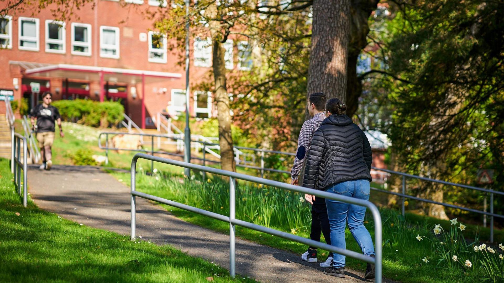 Students walk through the campus at UHI Perth. There is a red brick building and a path lined by trees.