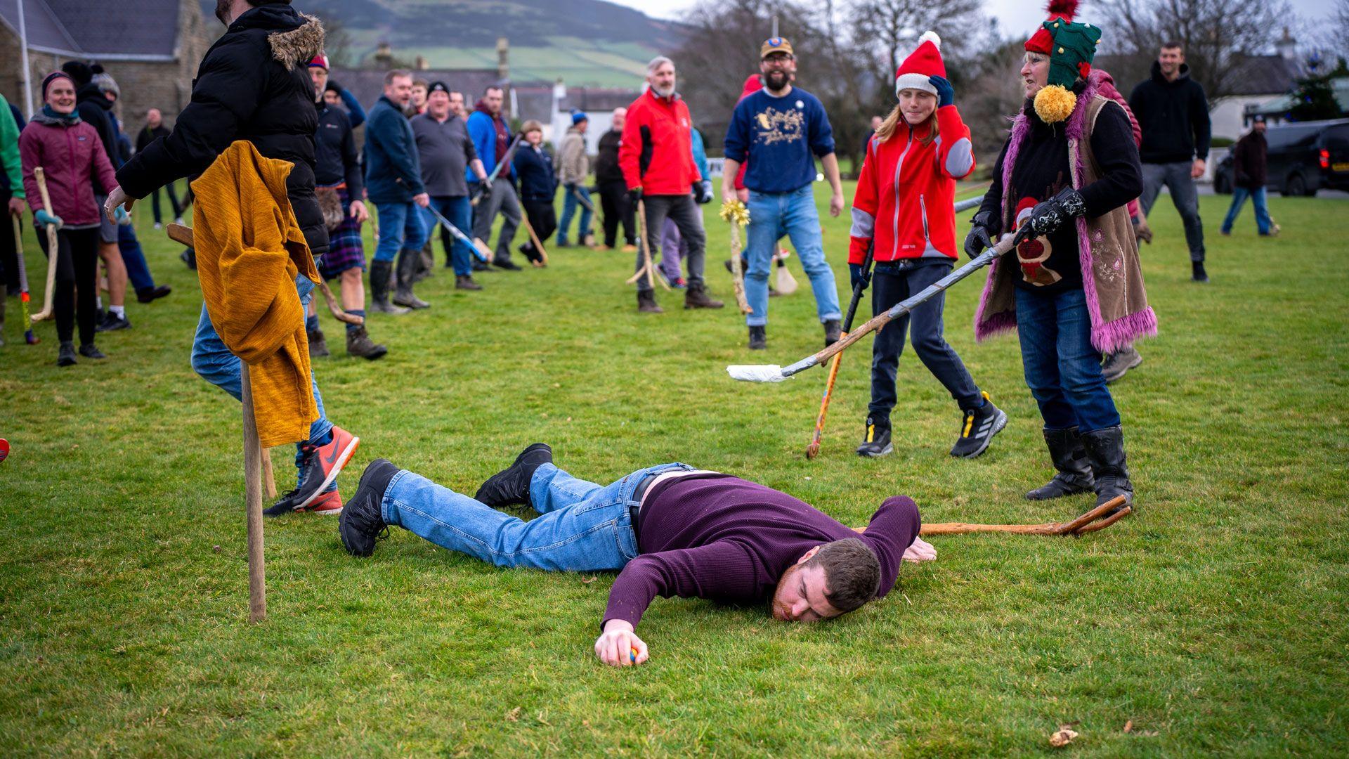 A goal keeper lies face down on the ground next to a goalpost while others with sticks look on.