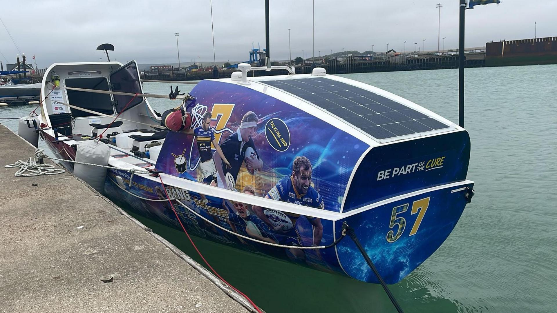 A close-up image of a four-person ocean rowing vessel moored in Newhaven Marina with bright coloured pictures of Doddie Weir and Rob Burrow playing rugby on the sides.