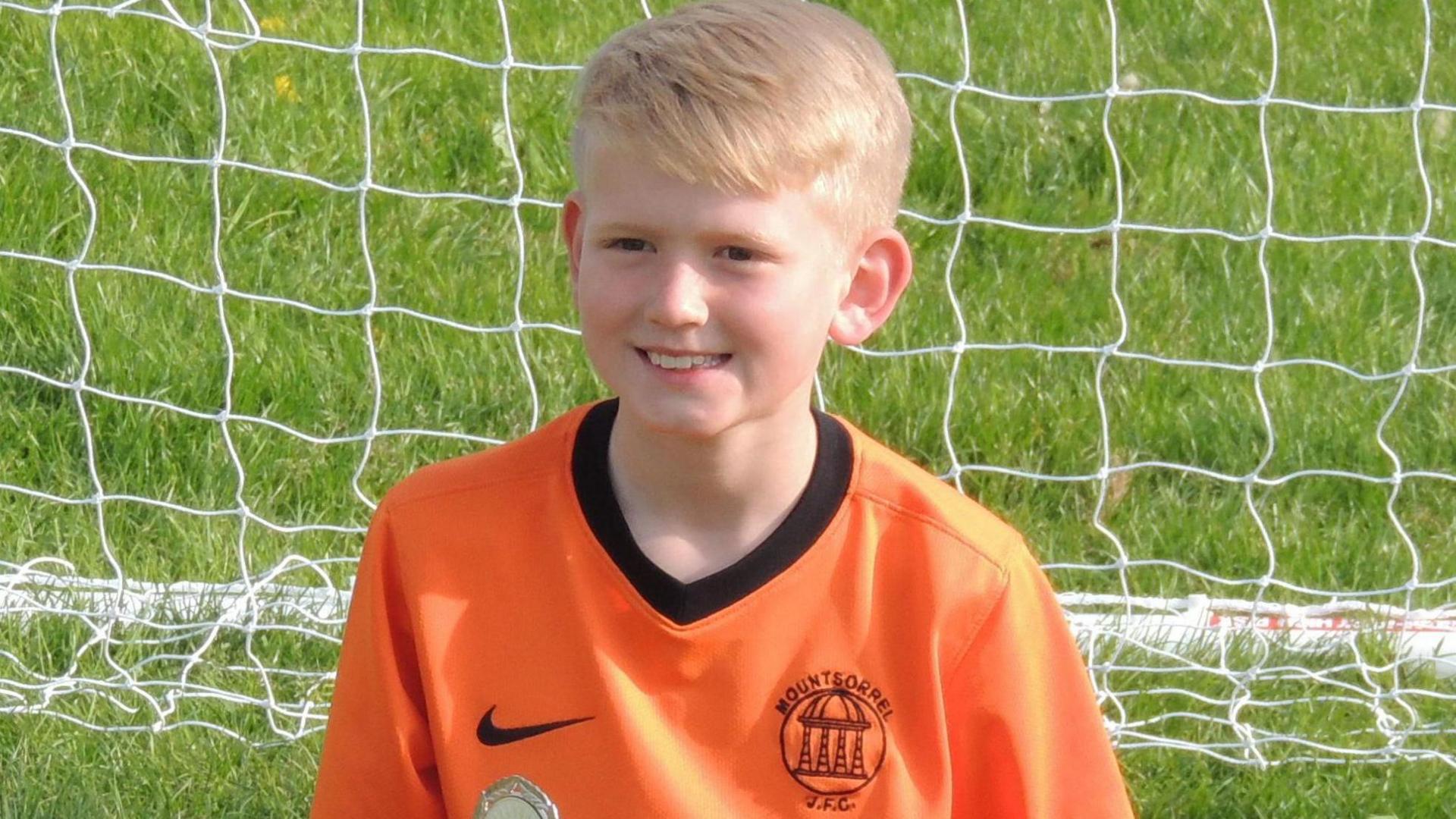 Sam Priestley smiling at the camera while wearing a Mountsorrel football shirt with a goal behind him.