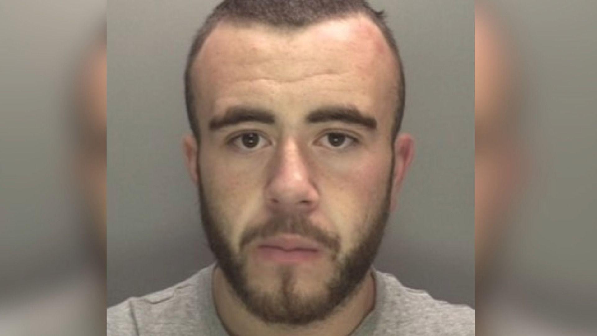 A police mugshot of Wayne Burke. He has short brown hair and a beard. He is wearing a grey top, and is stood against a pale backdrop.