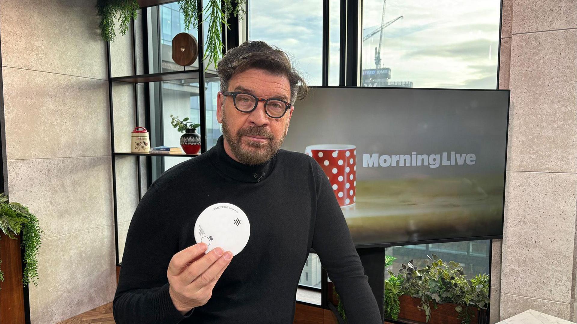 DIY SOS' Nick Knowles, sat in the Morning Live Studio with a brown shirt and straight facial expression. He is holding a smoke alarm.
