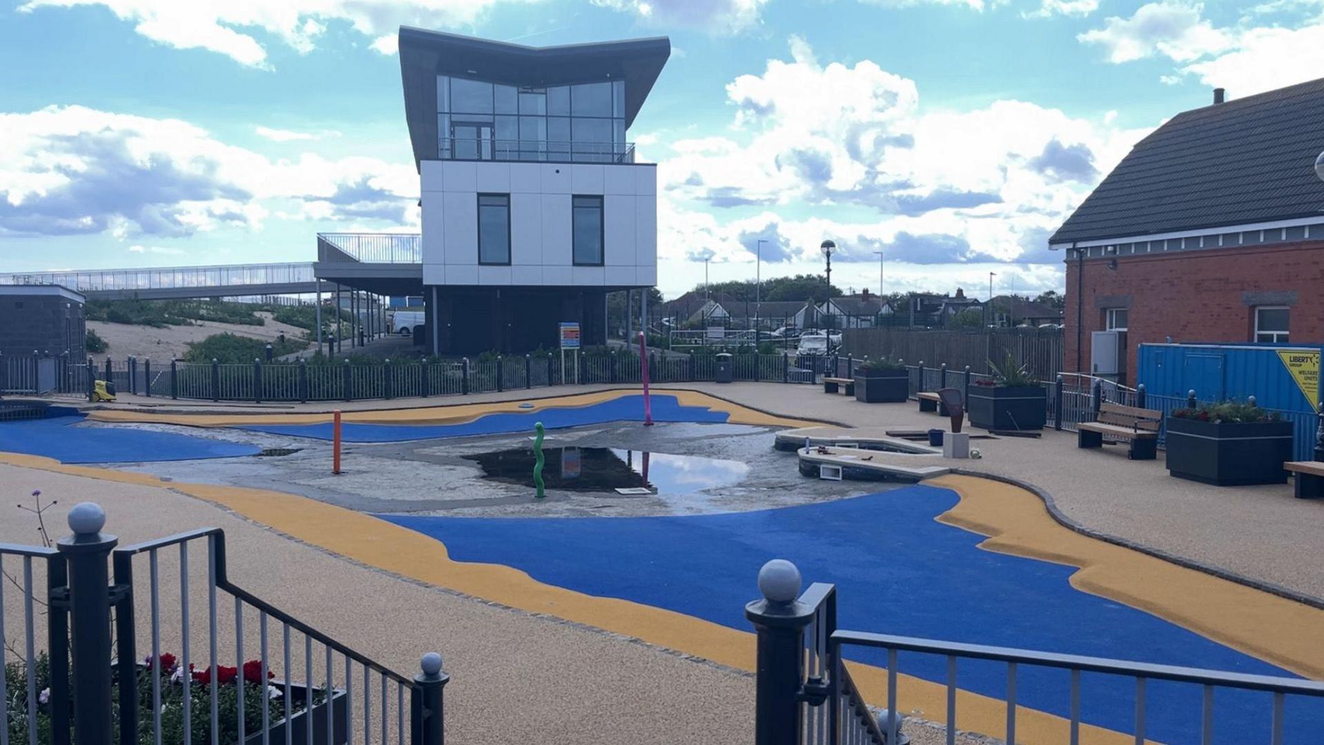 A largely empty paddling pool with a small amount of water in the bed. The floor is electric blue with a yellow border. In the background is a modern building dominated by a glass frontage.