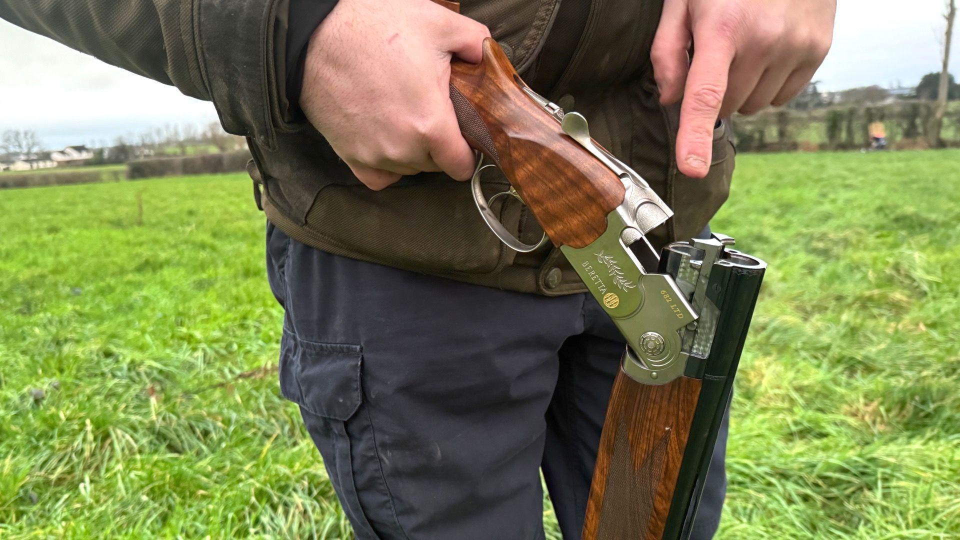 Picture of a man holding a shotgun.
The picture is cropped and you can only see the man from the waist to mid thigh. He is wearing a khaki jacket and navy trousers.
The chamber of the gun is open and the man is pointing to where the bullets go.