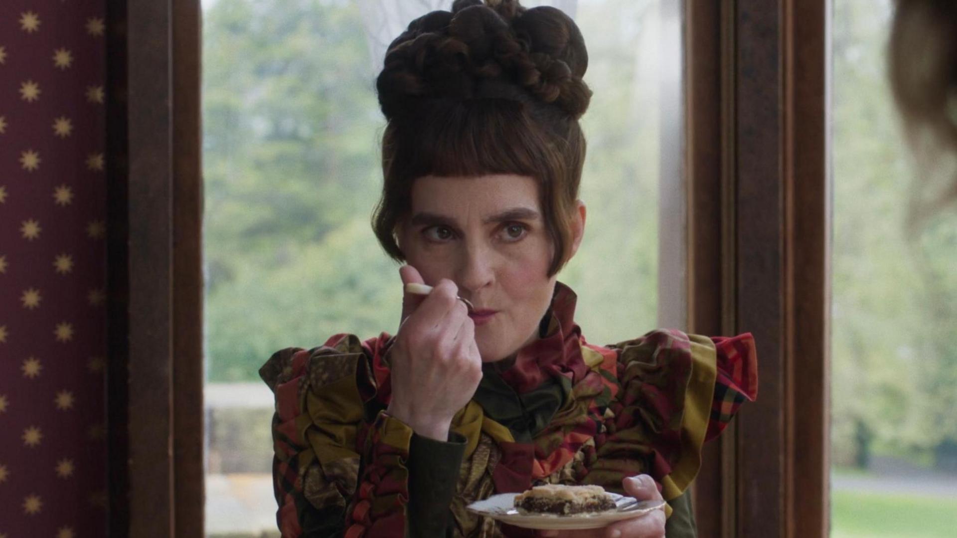 Actress Shirley Henderson is dressed in a period costume. She has dark hair which is styled on top of her head. She is holding a slice of cake on a saucer and taking a bite of it from a fork. She is sitting in front of two windows and greenery from a garden can be seen in the background. 