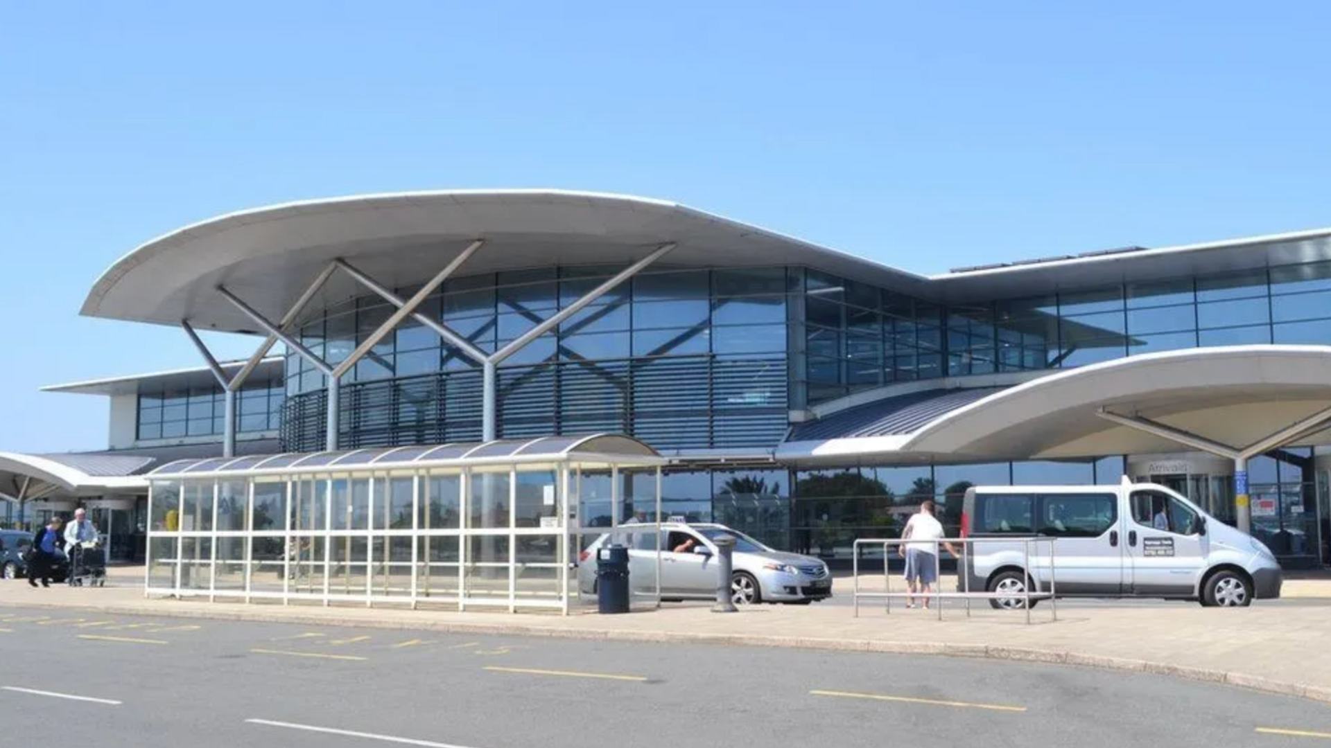 Guernsey airport - a building made predominantly out of glass windows, a taxi rank in front with two taxis, a road in front of it on a sunny day