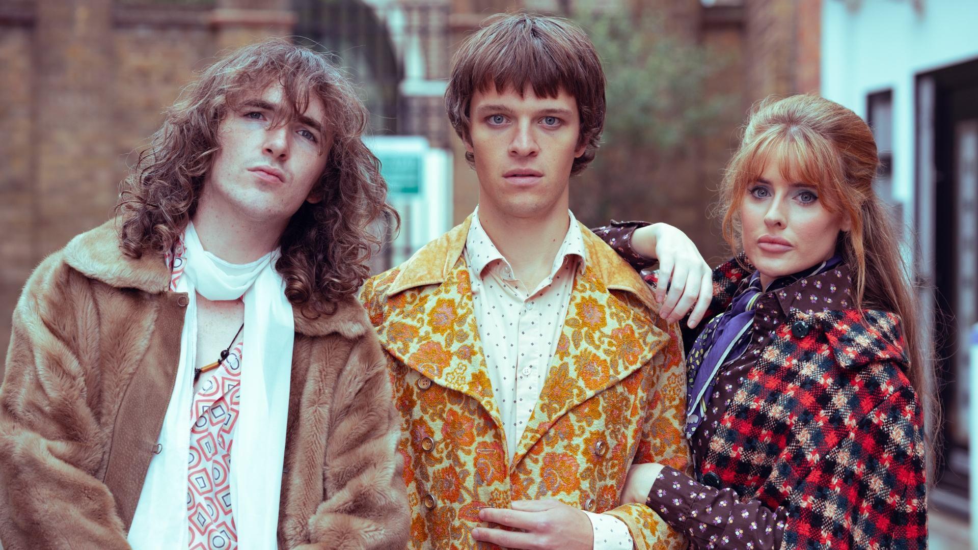 Three people. The white man on the left has a brown furry coat on and a while scarf, he has long, brown, curly hair. The white man in the middle has a floral orange and yellow blazer on over a white button-up shirt with small dark spots on it. He has a medium-length bowl cut. The white woman on the right had long light brown hair with a fringe. She is wearing a red and black tartan patterned overcoat. 