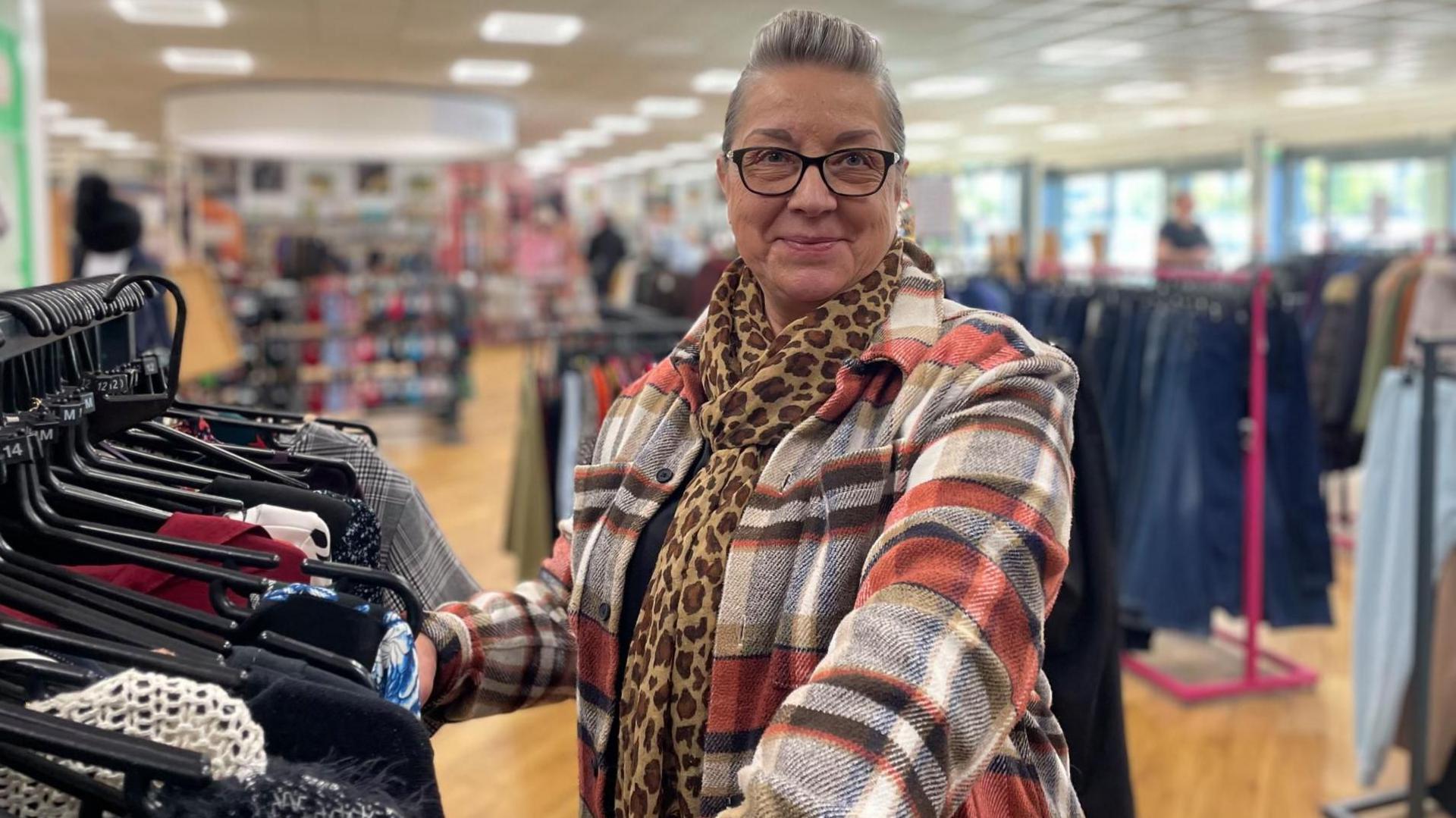 A woman in a colourful large checked coat and leopard-print scarf smiles for the camera in a charity shop.