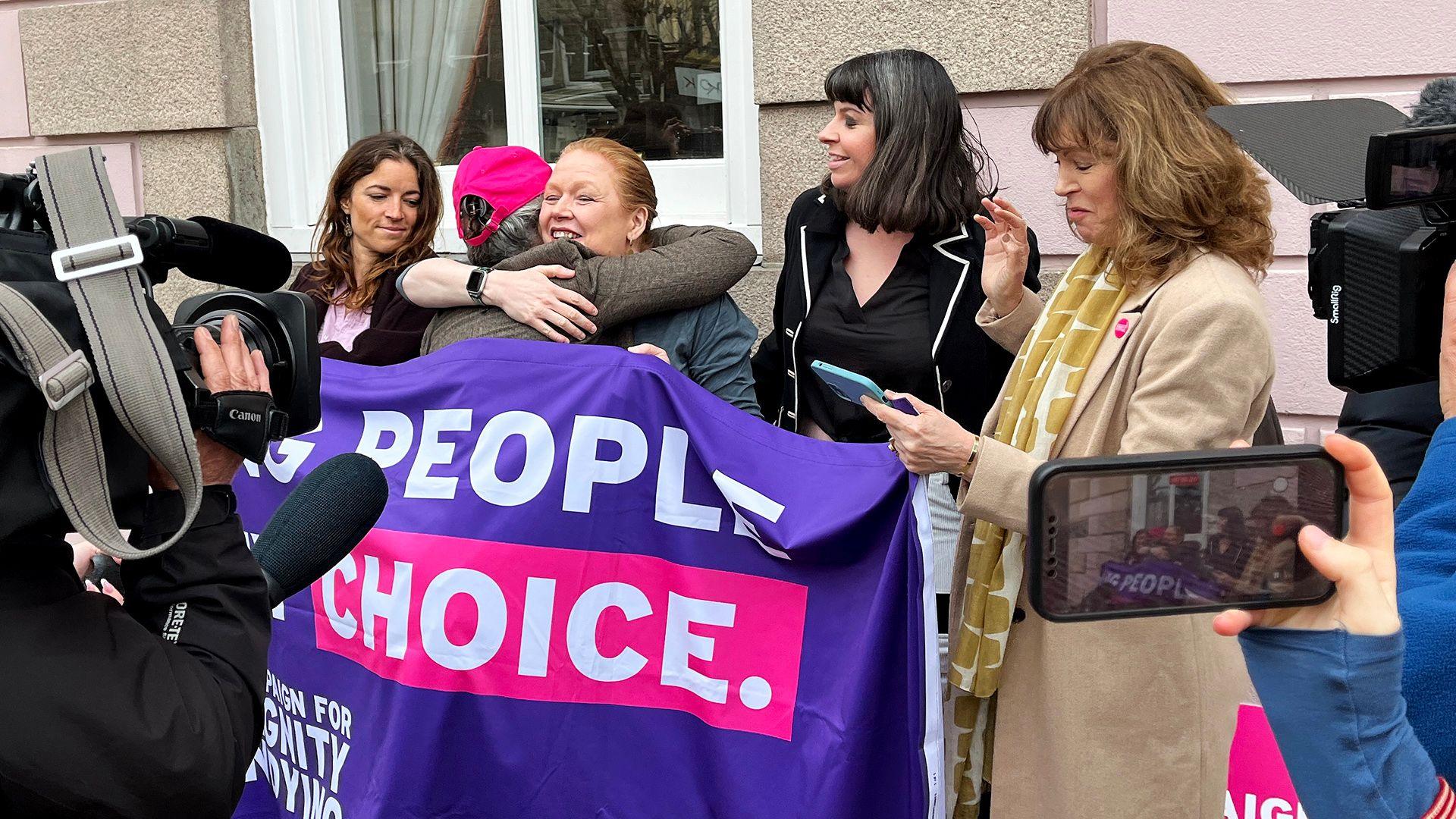 Five women celebrate with hugs next to a banner saying 'people, choice' with news cameras around them, outside on the street in Jersey on Thursday.