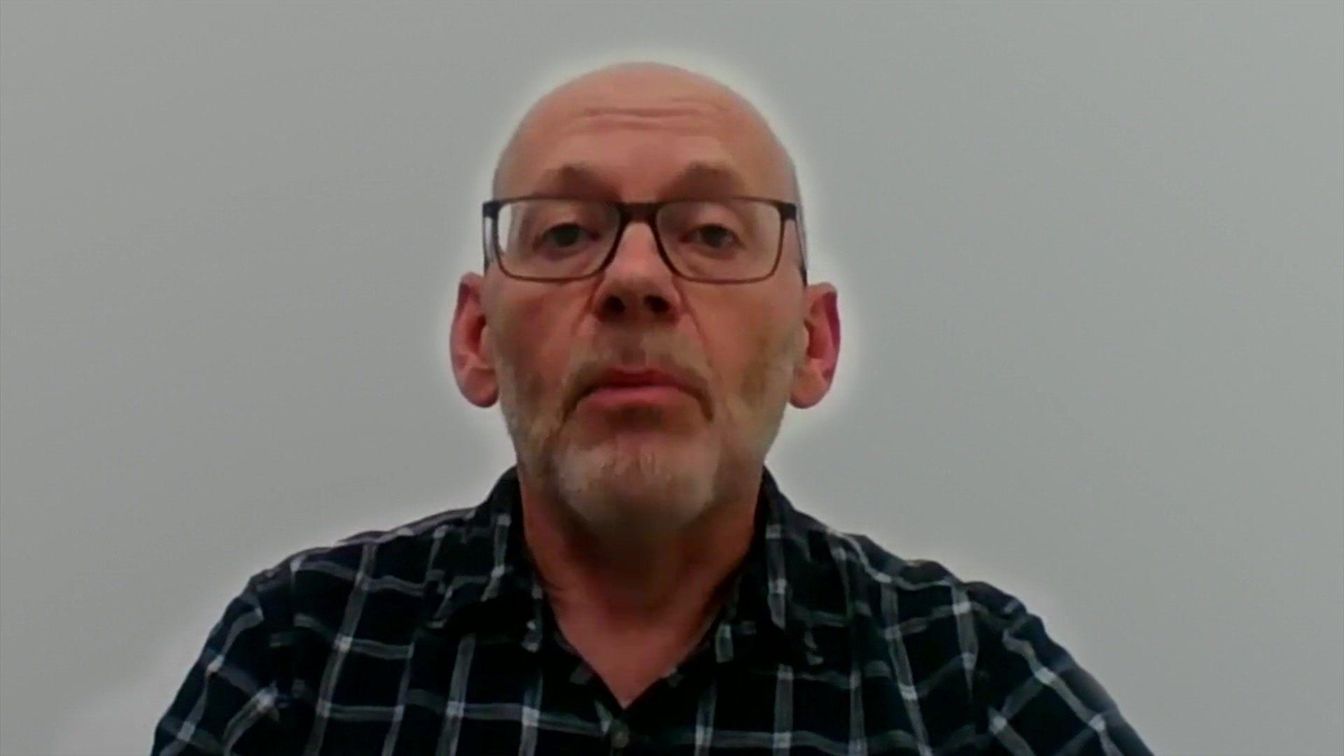 Peter Hambly in an interview. He is sitting down and facing the camera in a room, against a while wall