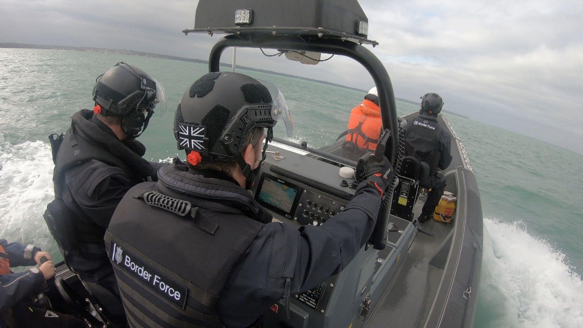 Uniformed Border Force officers, wearing helmets and stab vests, on board a fast inflatable RHIB vessel, powering through the water with wake and waves at its side. 