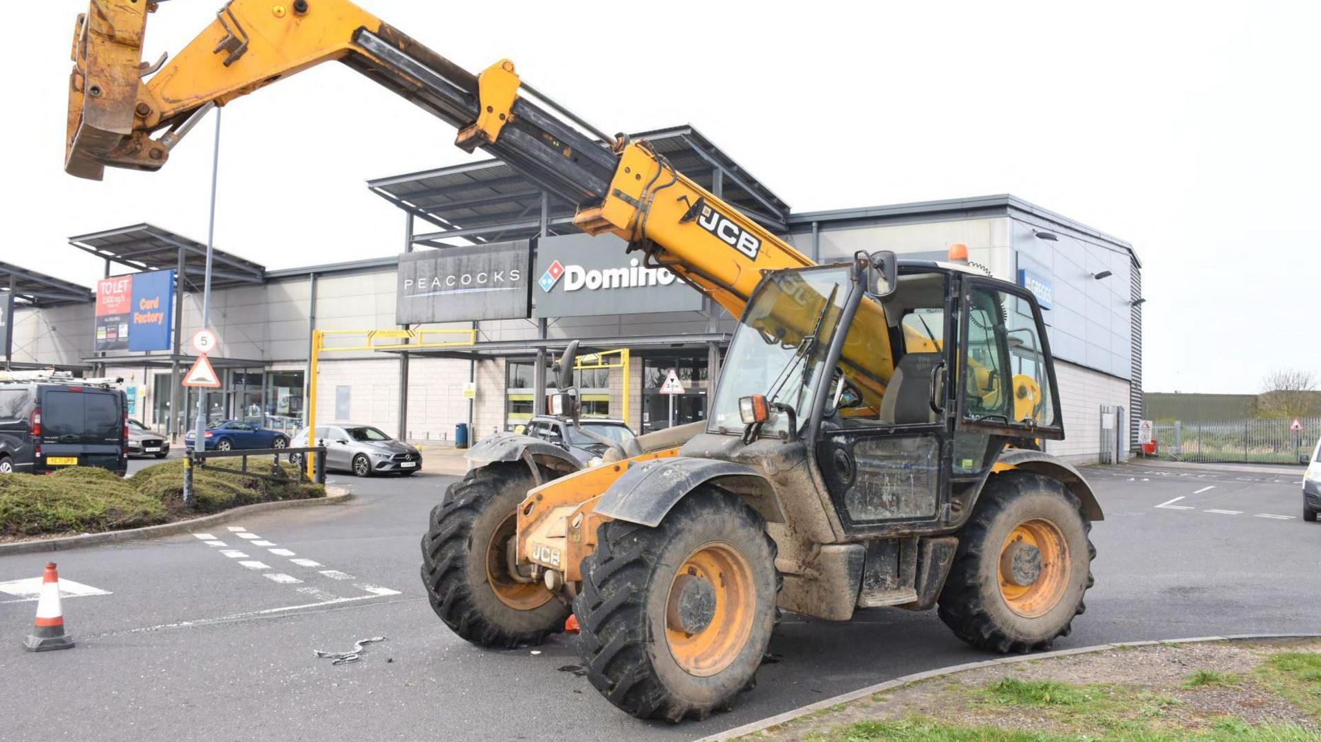 A large yellow and black JCB forklift truck left in front of shops at a retail park.