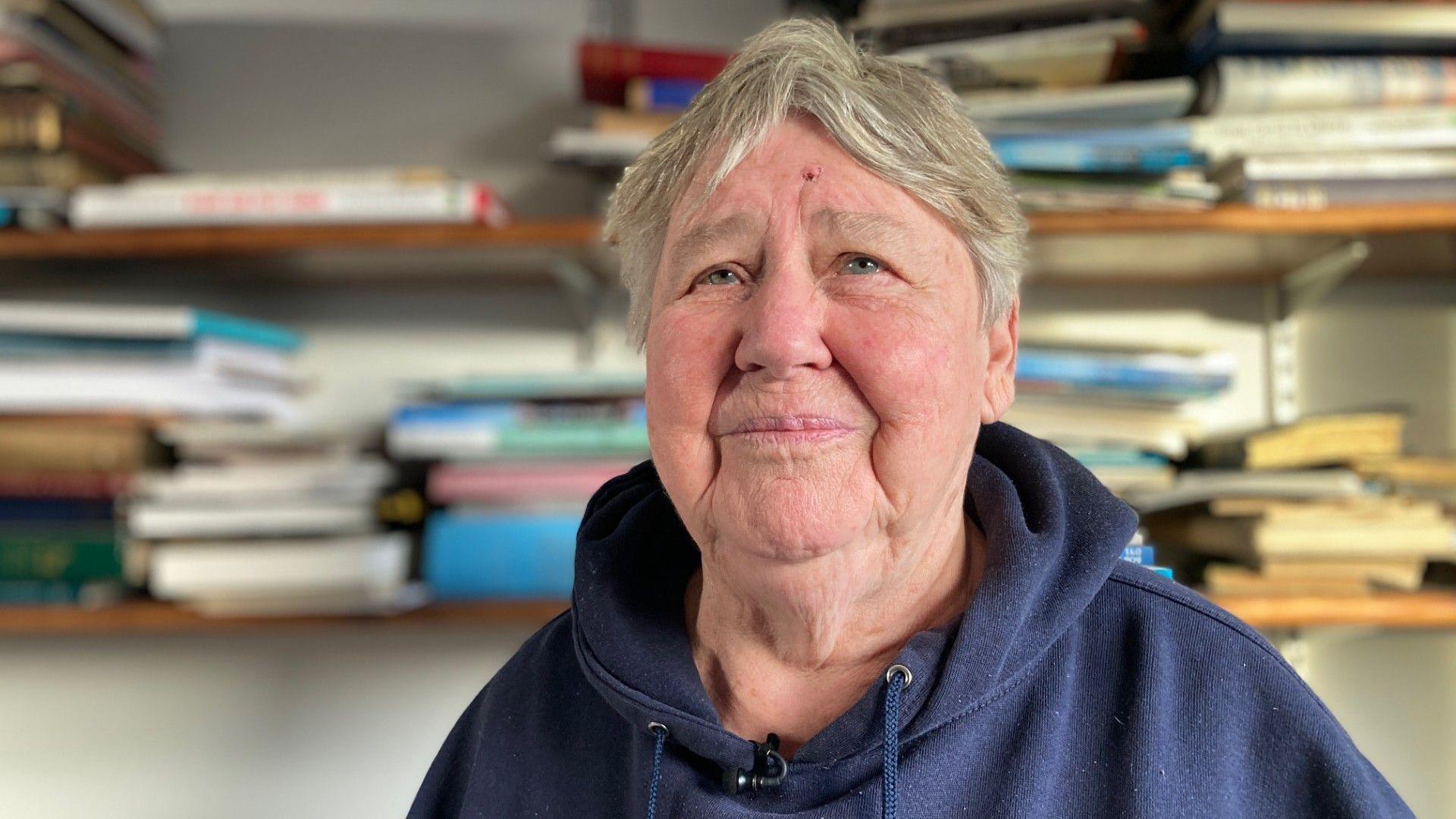 Maragaret Jones yn sefyll o flaen silff lyfrau