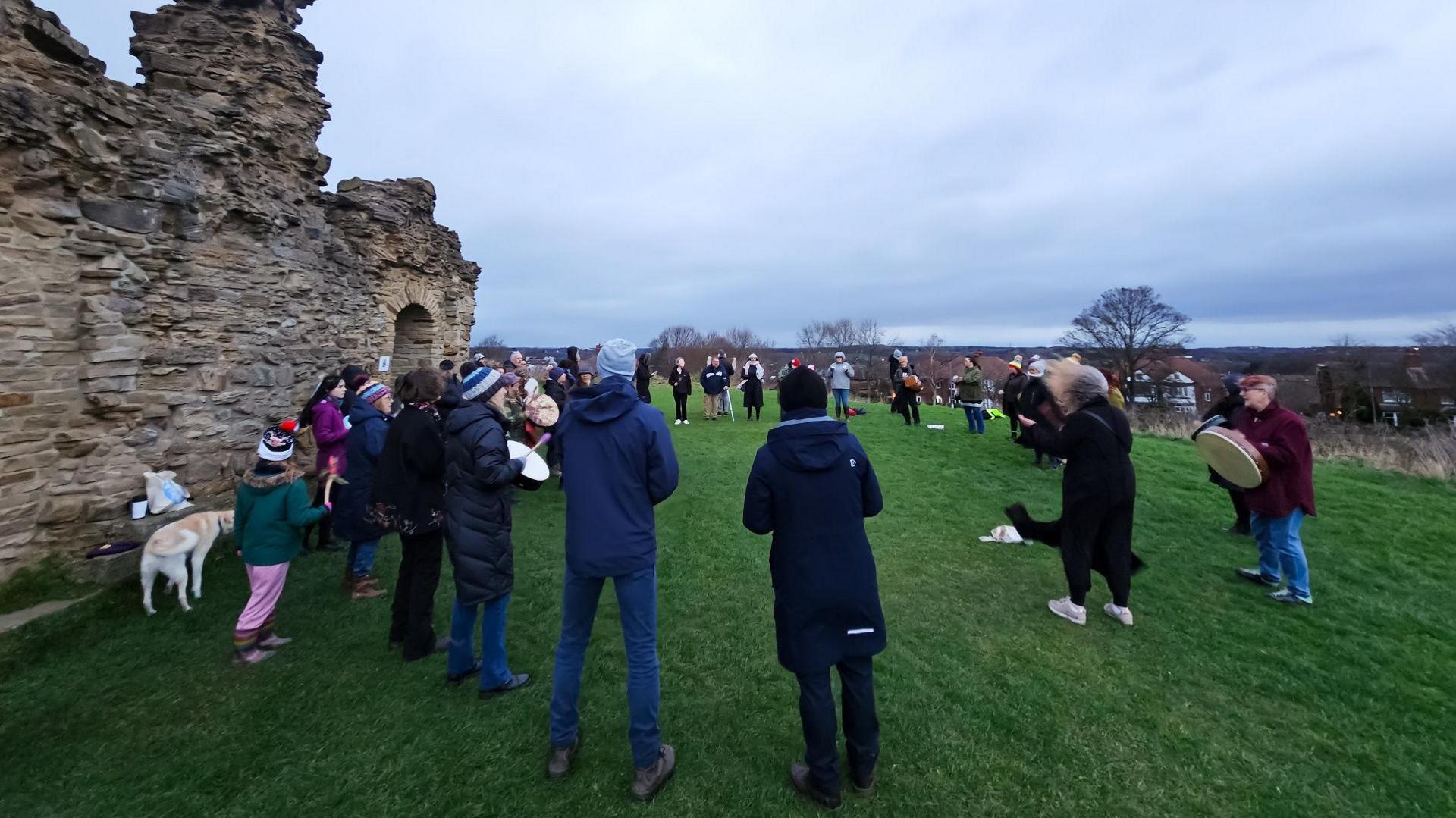 More than 20 people stand in a circle near a ruined castle drumming. They are joined by one dog