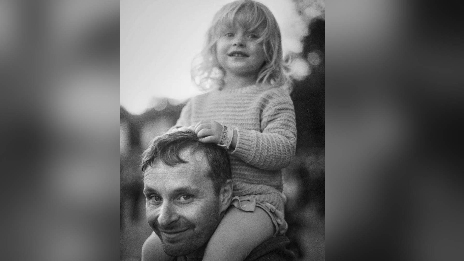 Stu Housley pictured with his young daughter, Elin, on his shoulders. She grips on to his hair with her hands and he smiles at the camera. She has blonde hair in a full fringe and curls and smiles at the camera. The image is black and white.