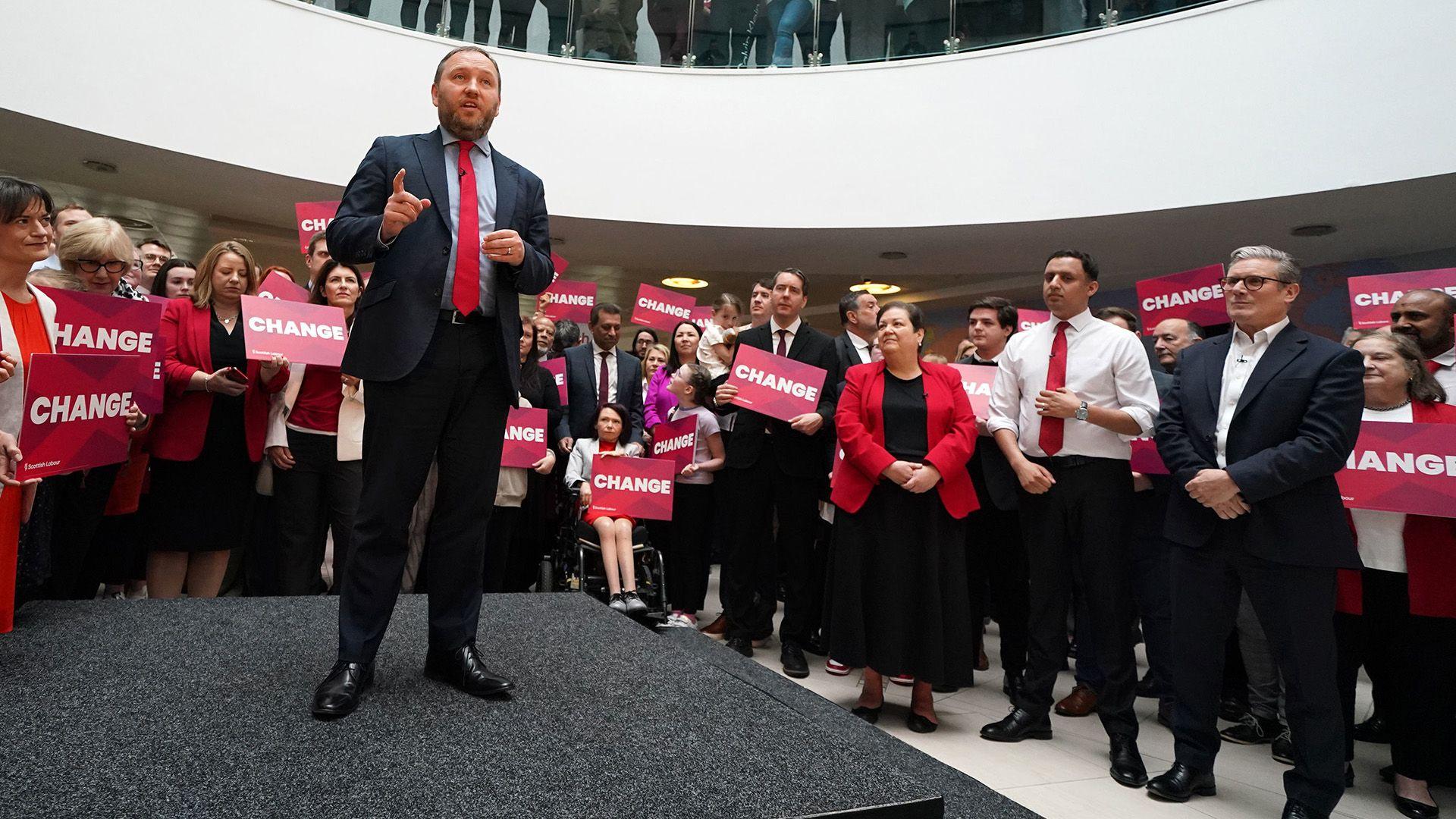 Ian Murray stands on stage in a suit and red tie and men and women in suits and smart clothes hold red signs saying change