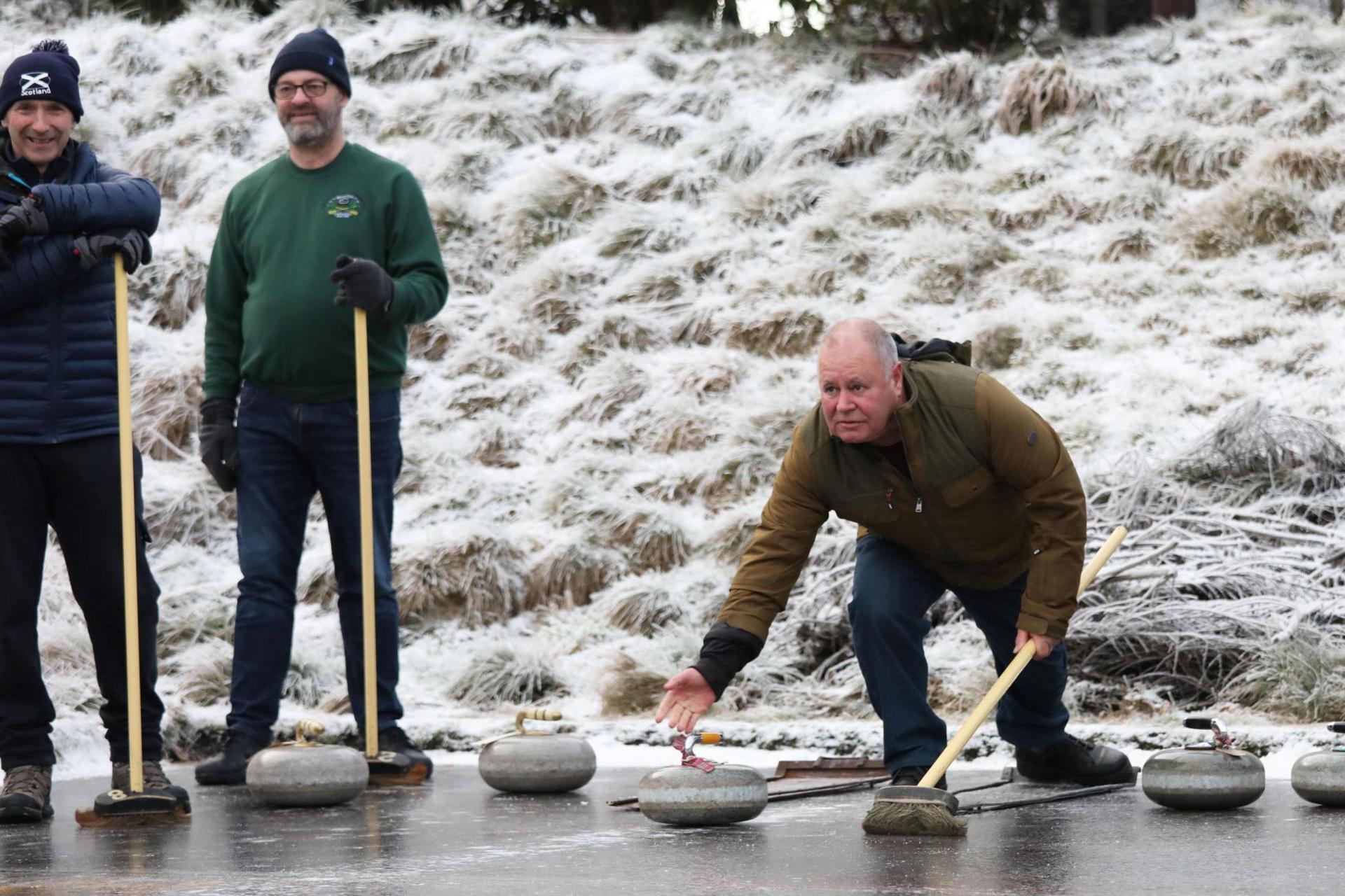 Three curlers stand on the edge of the rink. One of the curlers is leaning over and pushing a stone out on to the rink.