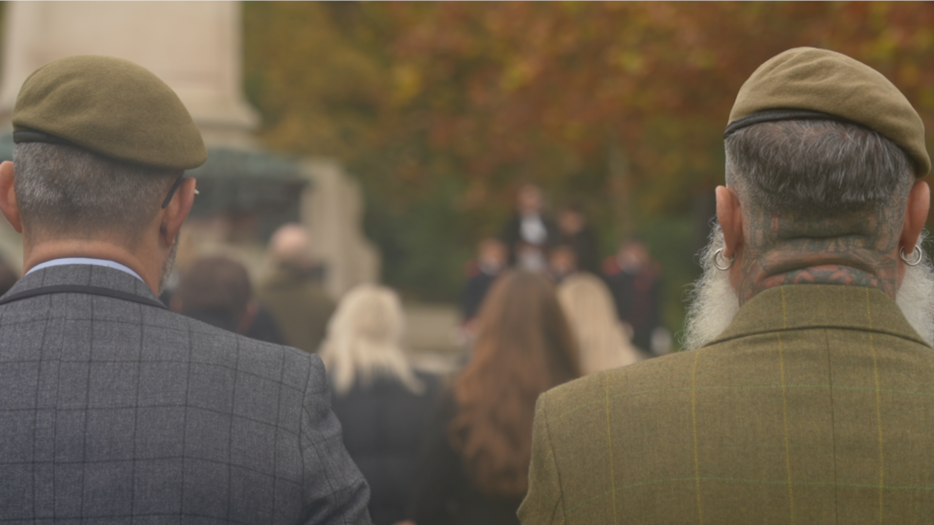 Two men with their backs to the camera at the cenotaph in Christchuch Park in Ipswich. They are both wearing green berets and checked jackets, one grey the other green. Beyond them, a service can be seen taking place.