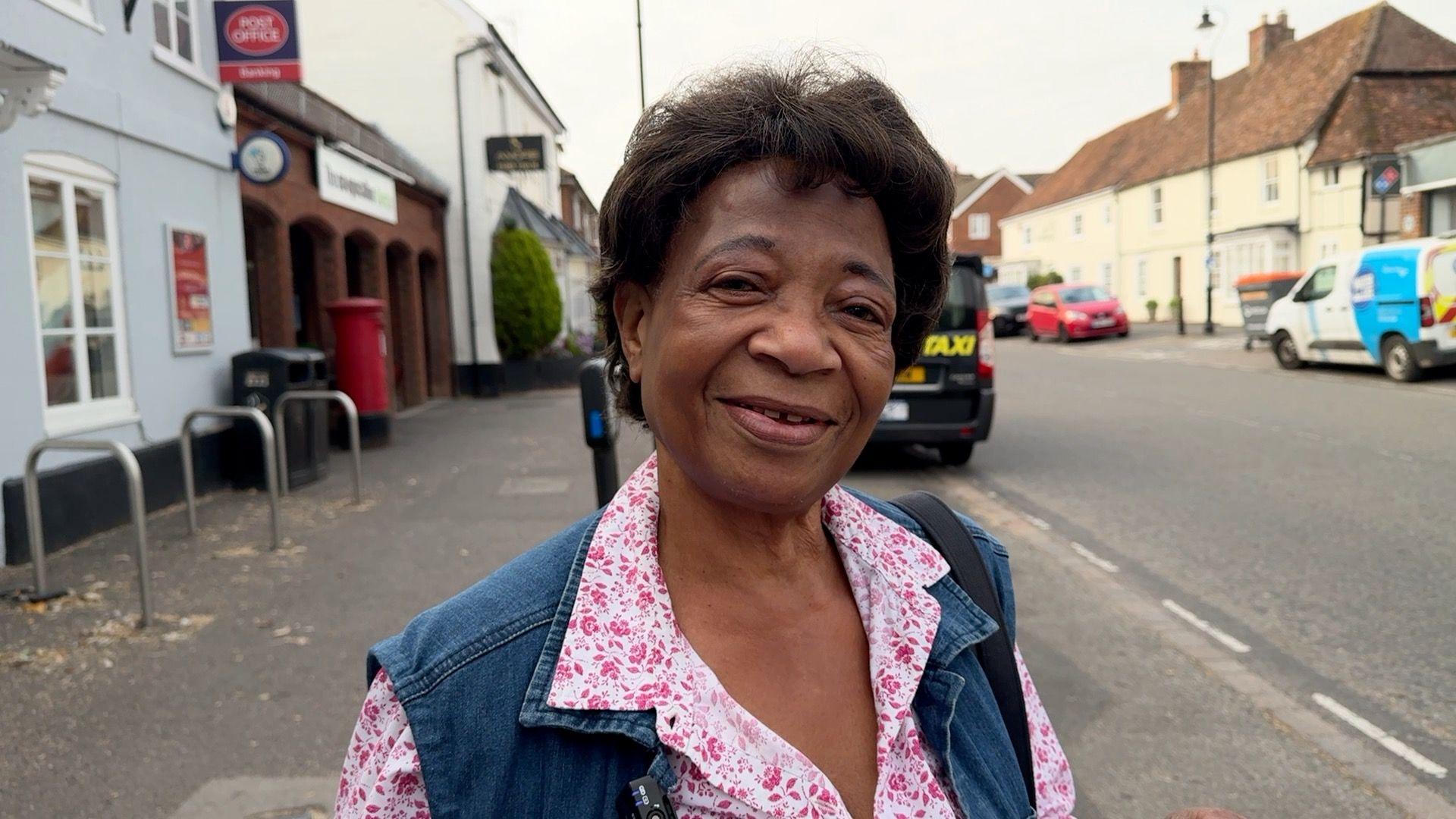 A woman with short dark brown hair, a pink floral-pattern blouse, and a denim waistcoat-style jacket, standing in a road and smiling.