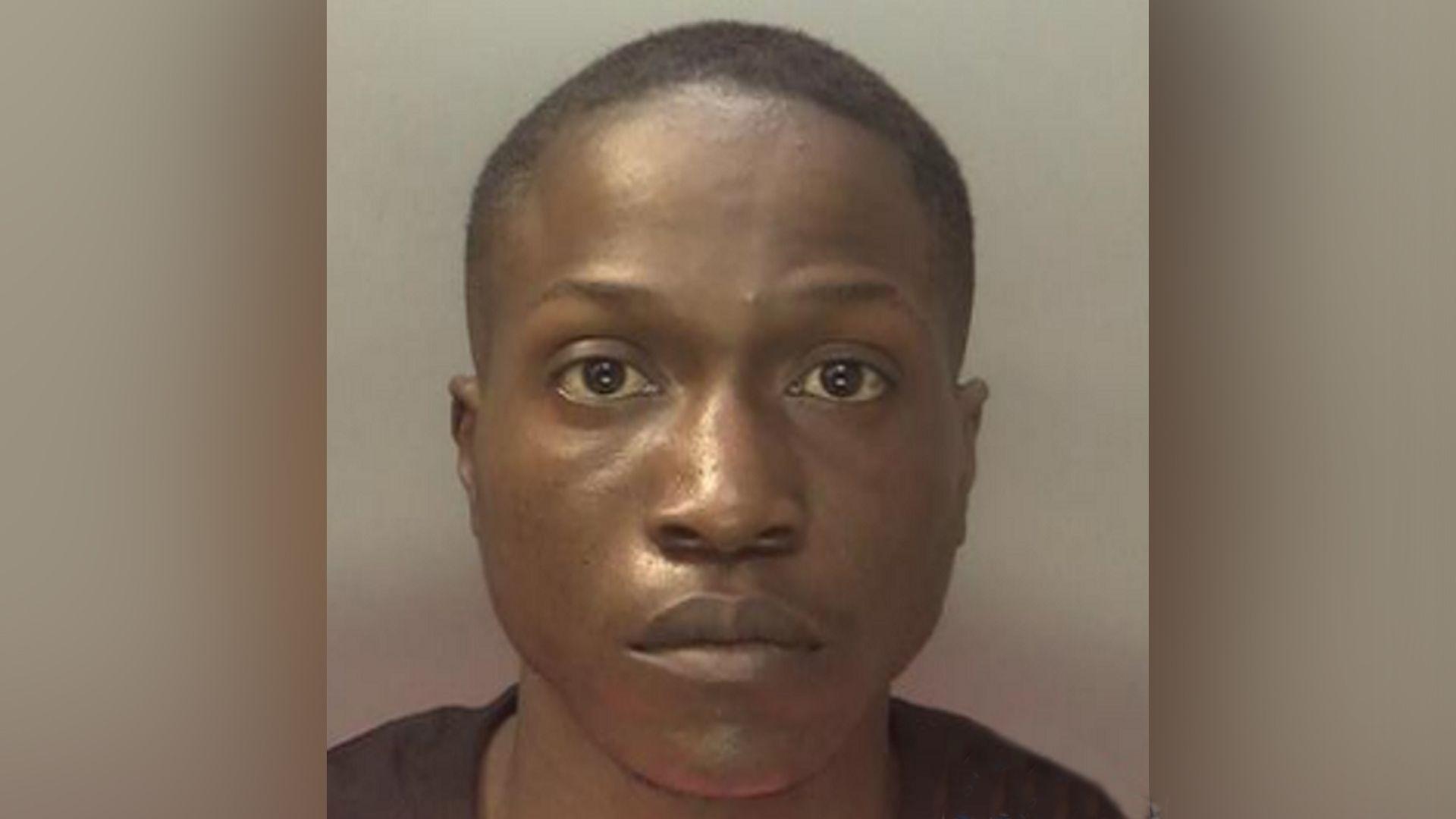 Police mugshot of a man with close-shaven black hair and wearing a black T-shirt. He is looking straight at the camera.