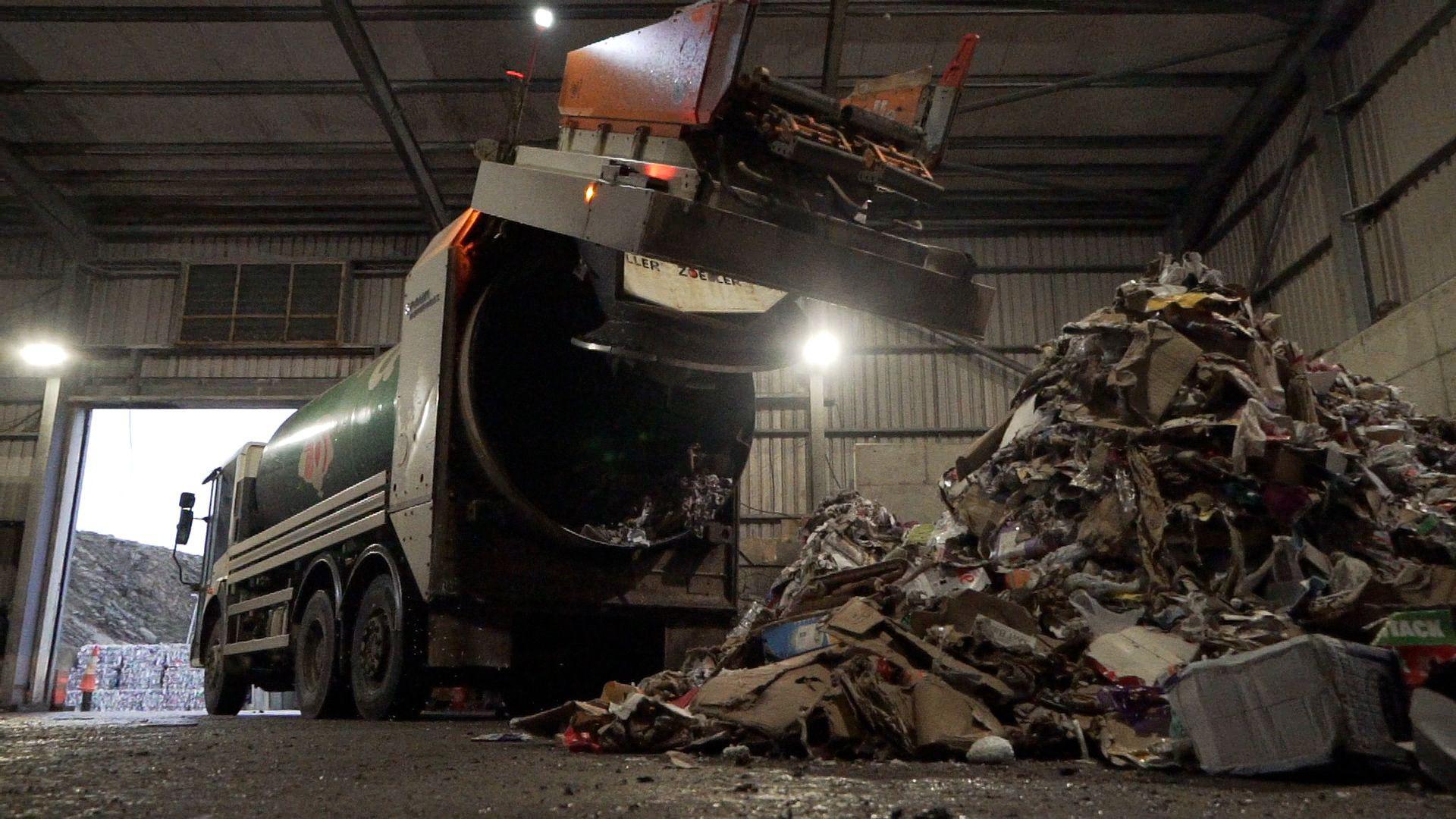 A bin lorry is emptying its load inside a facility built of corrugated metal. A large pile of waste can be seen deposited in the right hand side of the image.