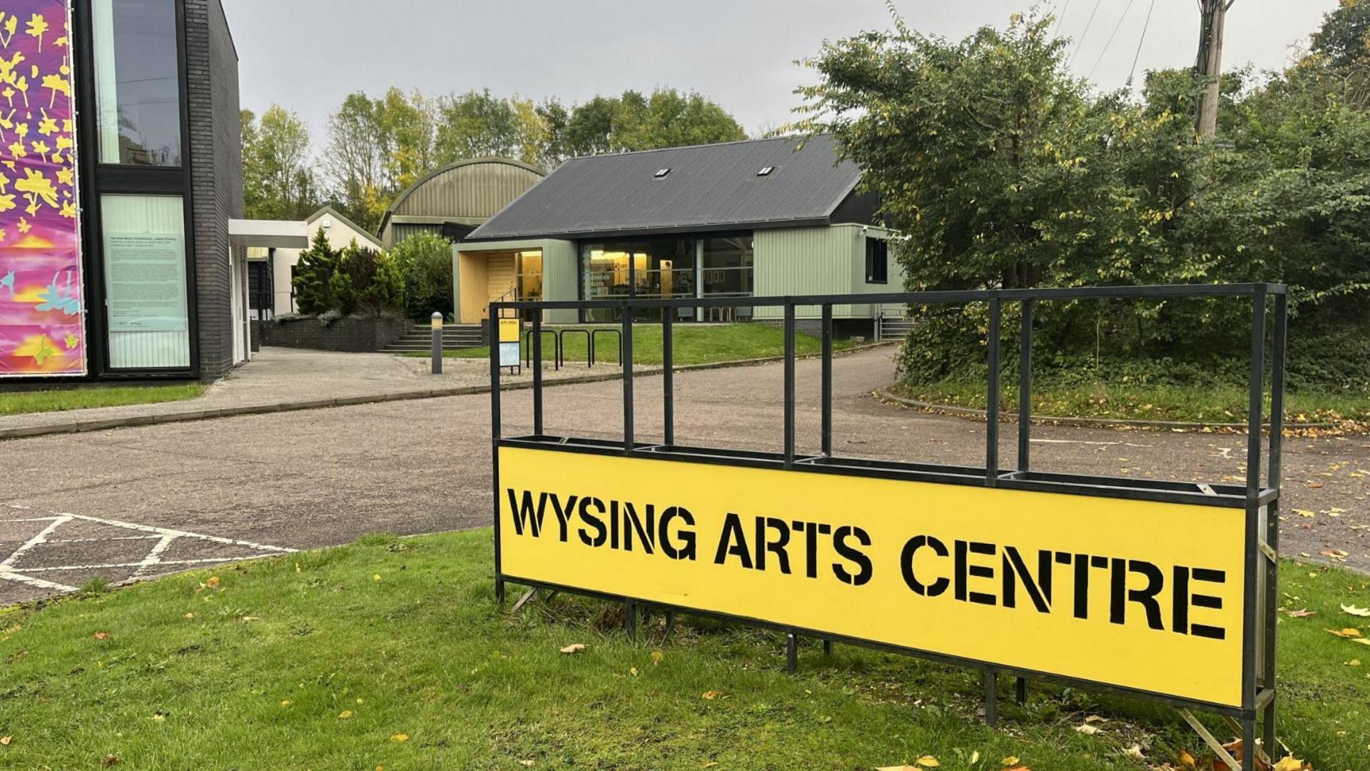 A yellow sign saying Wysing Arts Centre situated on grass with a green one-storey building in the background and a two-storey glass building to the left.