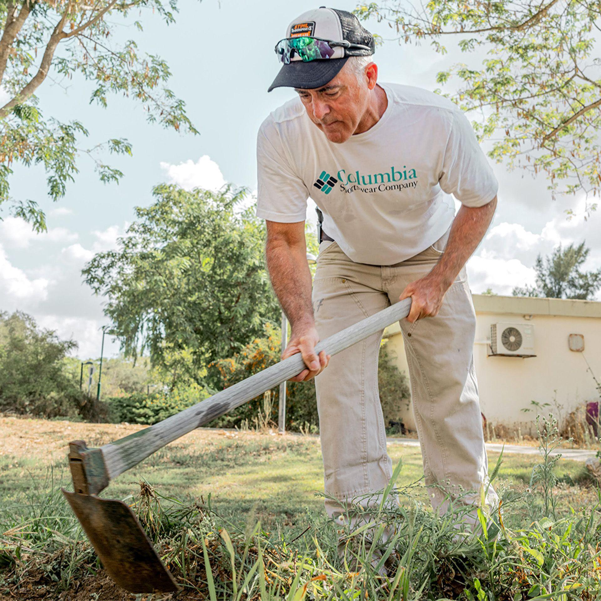 Simon King, in a white T-shirt, baseball hat and beige trousers, wields a gardening tool as he tidies the kibbutz gardens. He is surrounded by greenery.