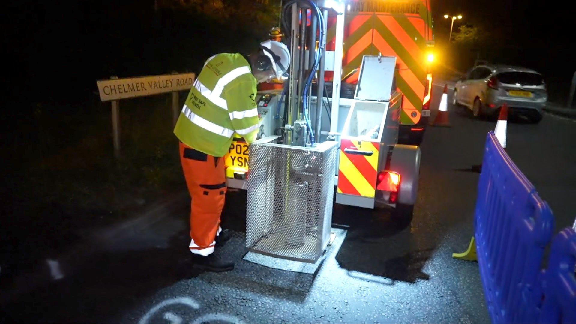 A highways van with flashing yellow lights is parked on a road. At the rear of it is a machine which contains a large drill which is lowered on to the road surface as it spins to remove circular samples from the road surface. It is night time, and so dark. A worker in orange trousers a yellow jacket is operating the machine. He is wearing goggles and a white hard hat.