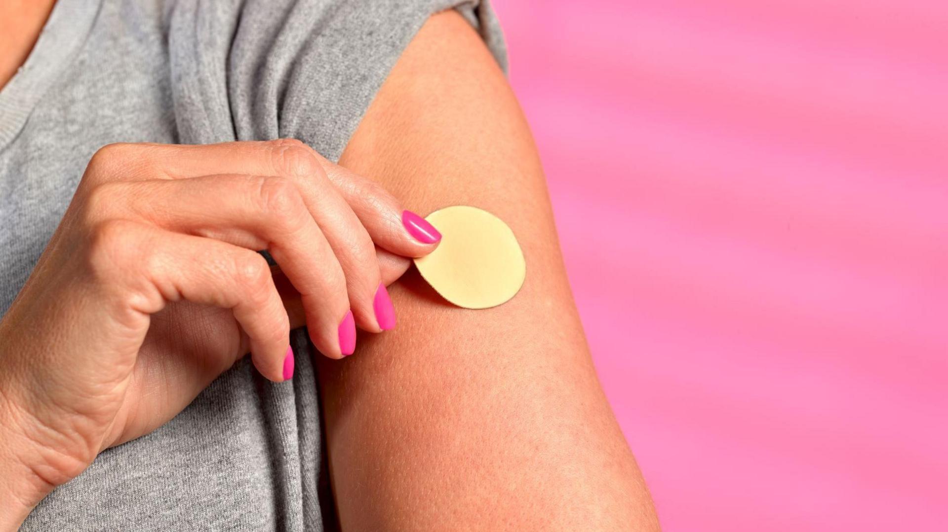 A woman places a nicotine patch on her left upper arm. She has painted, pink nails and wears a grey t-shirt. The background is pink.