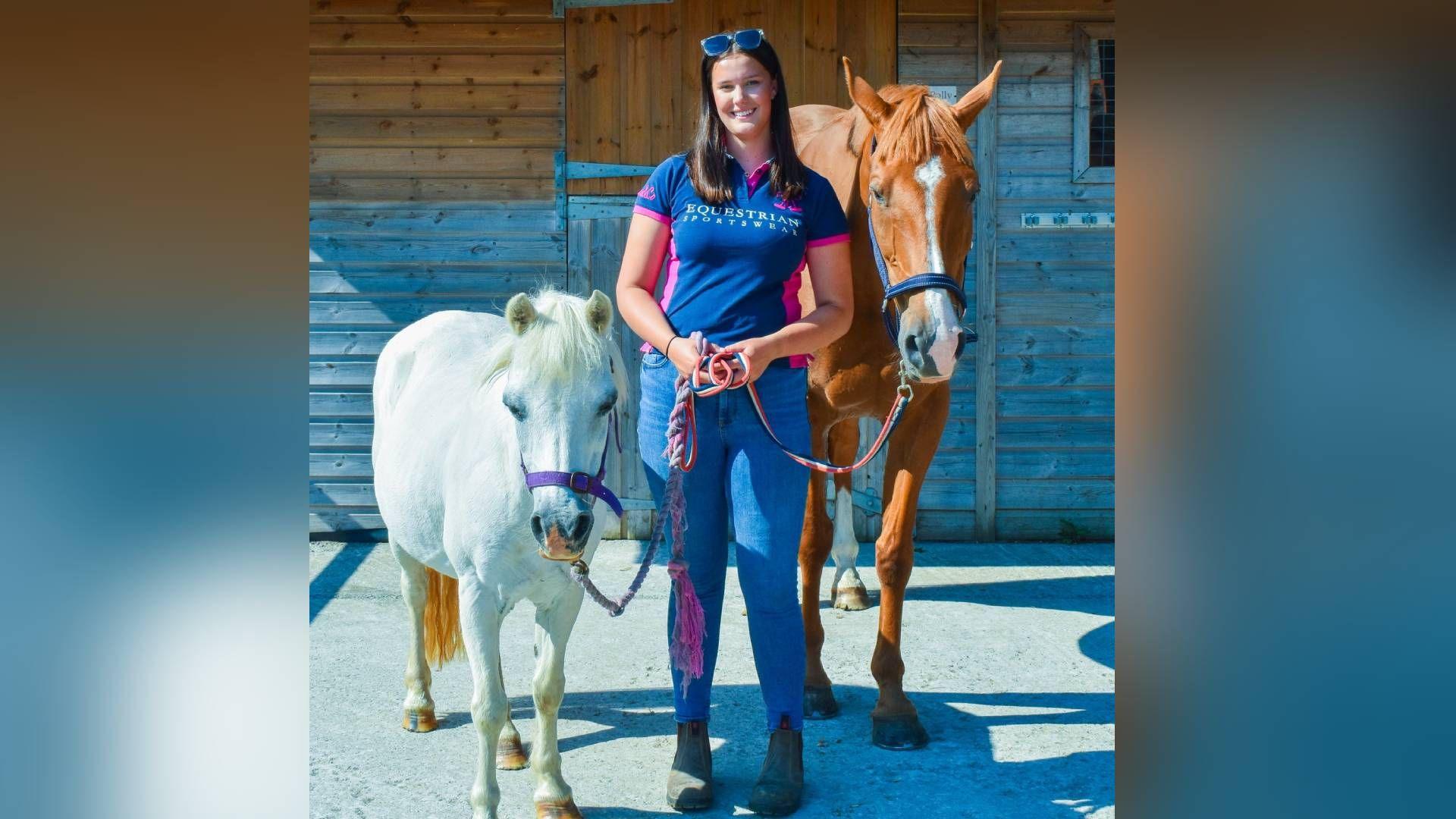 Sally Allen, 18, pictured stood side by side with two horses. She holds their reigns in her hands and smiles at the camera. She has brown long hair, with sunglasses on top of her head and wears chelsea boots, blue skinny jeans, and a blue polo shirt. 