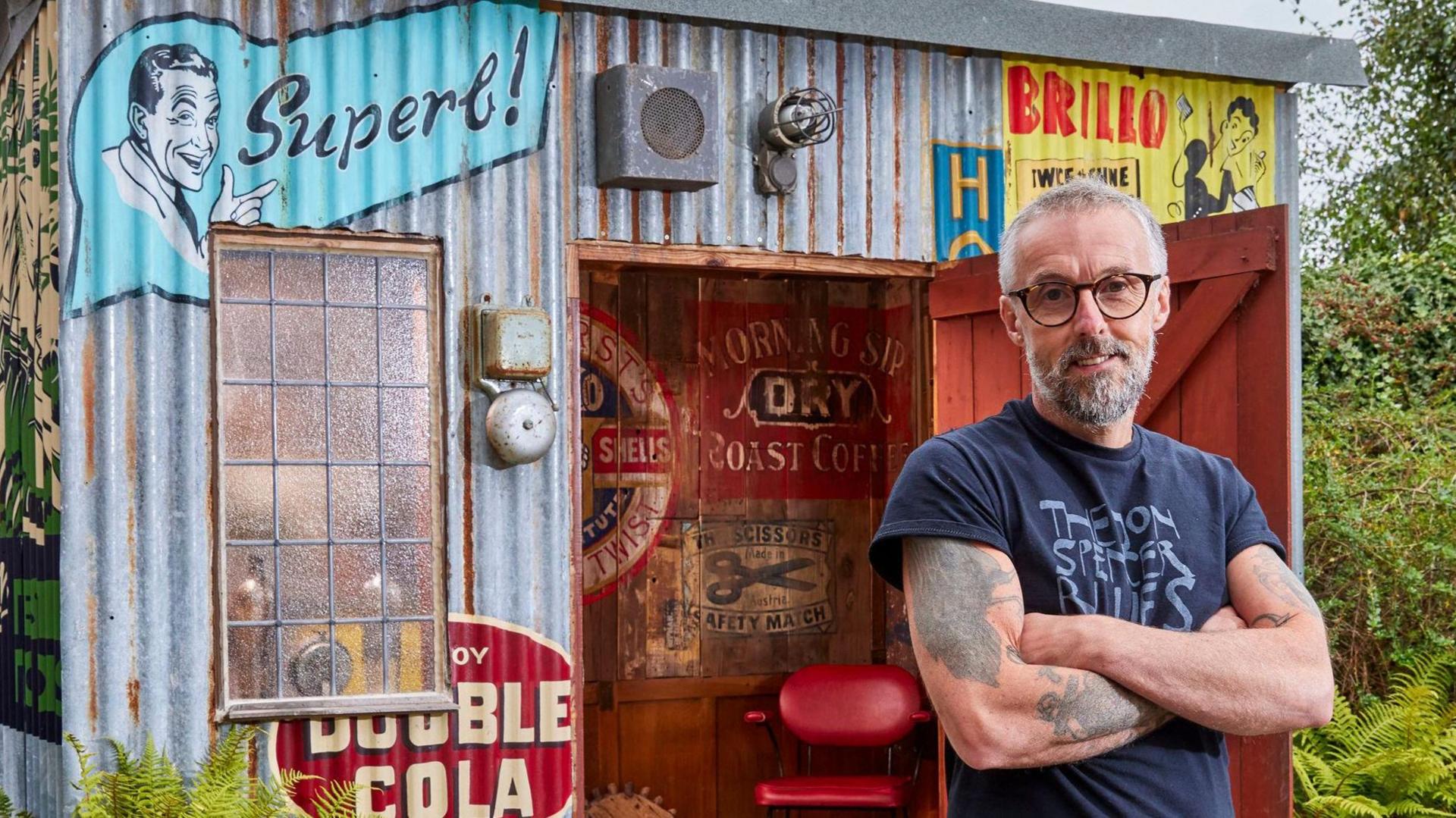 A man in a navy blue T-shirt and jeans stands in front of a corrugated shed with colourful painted slogans