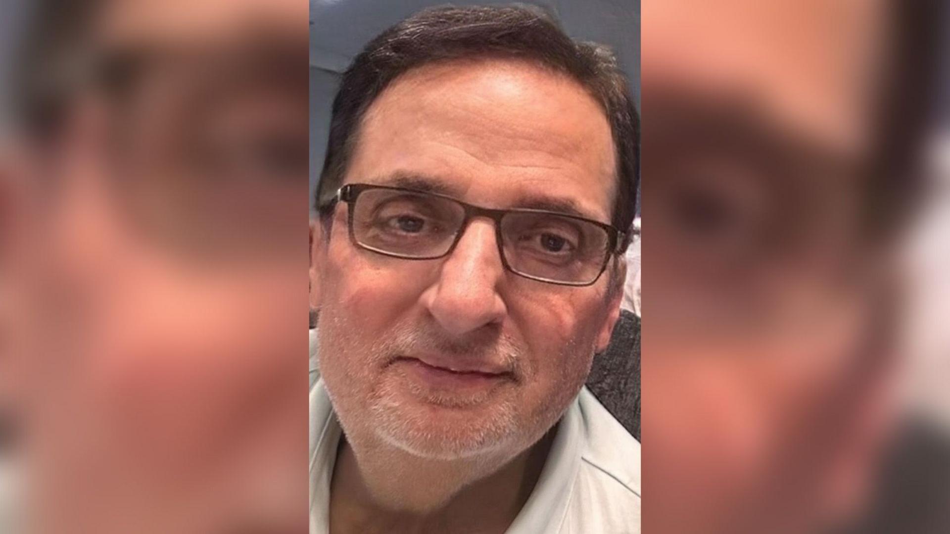 Rajinder Verma wearing glasses and a white shirt, with dark hair and a grey beard, leaning slightly to one side and looking at the camera.