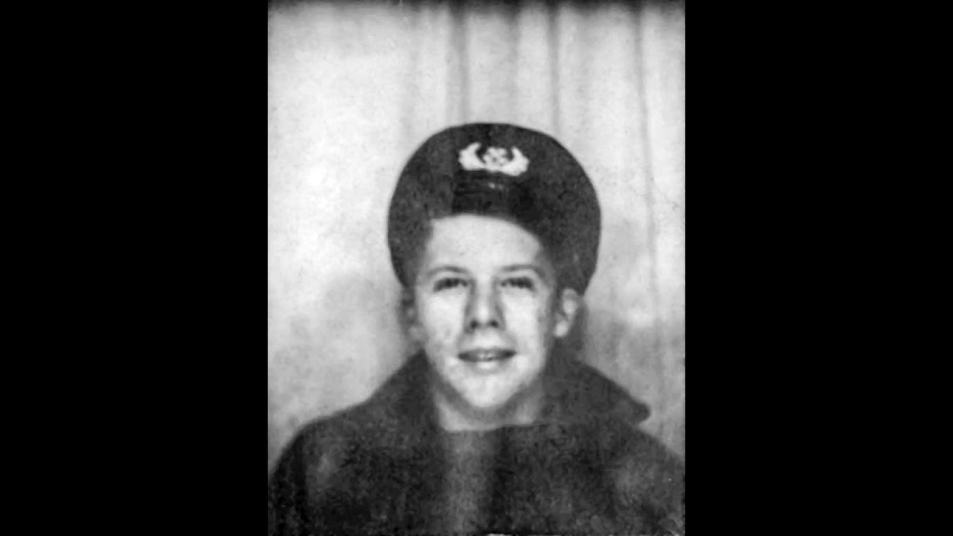  A black and white photo of a young man wearing a dark coat with a large collar and a cap with a US Navy badge on it. He is smiling.