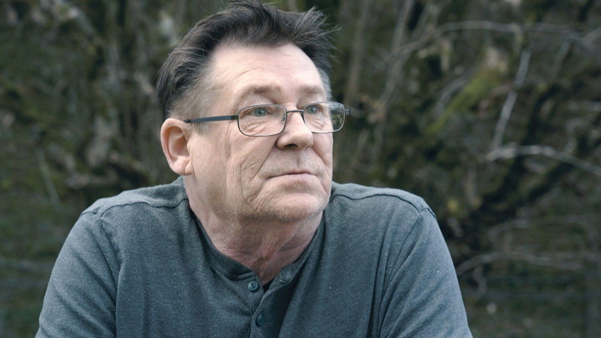 Martin Cullen, with dark hair and glasses, looks off to the right thoughtfully. He is wearing a grey button-through top and there are bare, wintery trees out of focus behind him.