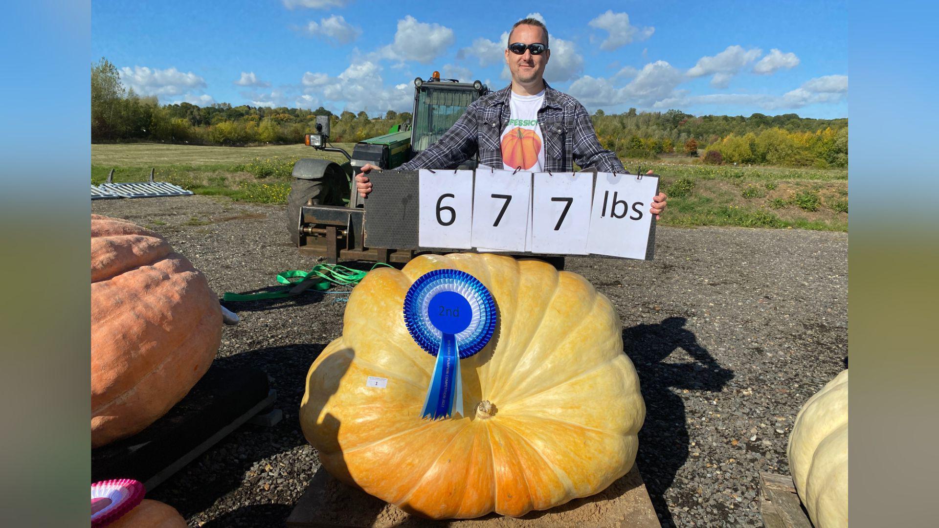 A man, who is wearing sunglasses, holding up a sign which reads 677 pounds. He is standing next to a giant light orange pumpkin.