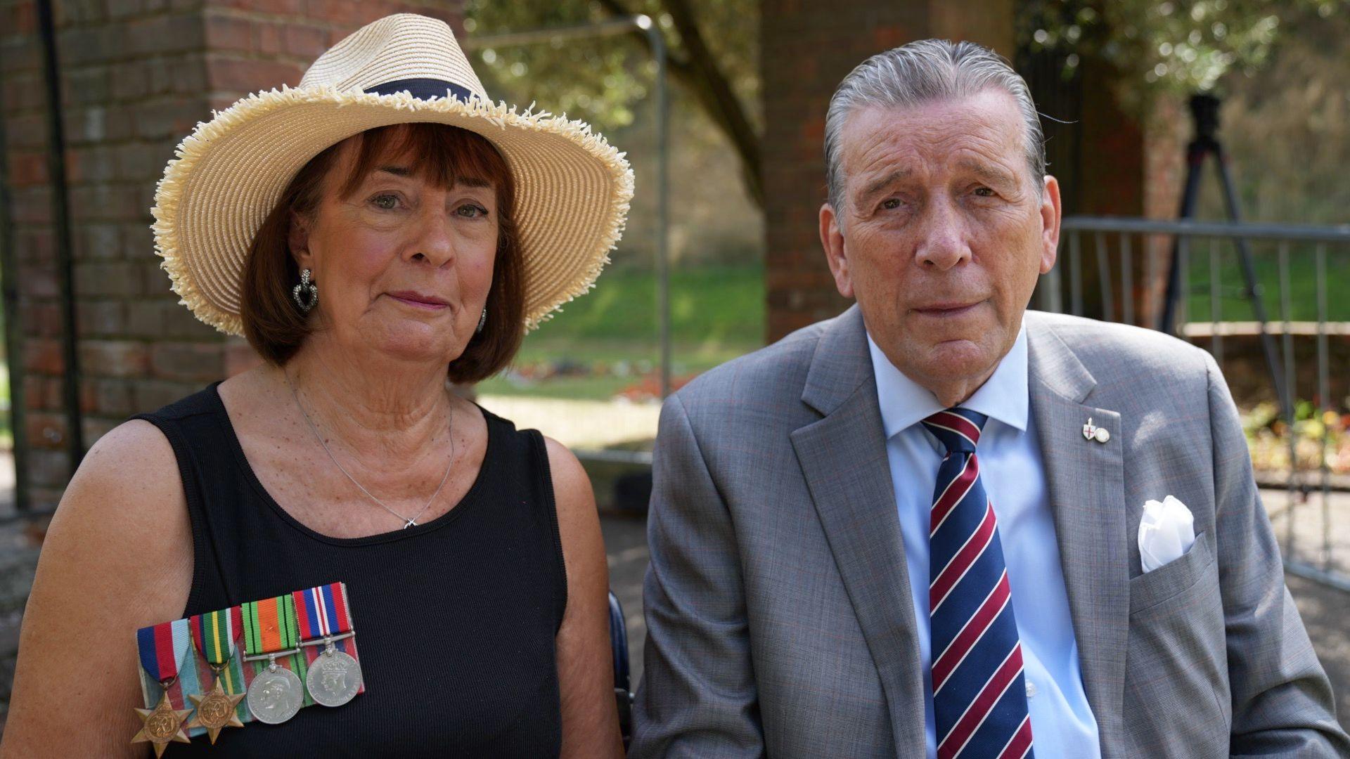 Gillian Powell sits with her husband Stephen on her left-hand side. She is wearing a black dress with her father's military medals attached. She is also wearing a straw hat and a silver necklace and earrings, and she has dark brown hair. Stephen is wearing a grey suit jacket with a light blue shirt and dark blue and red striped tie. He has a white handkerchief in his breast pocket, and has greying hair which is combed backwards.
