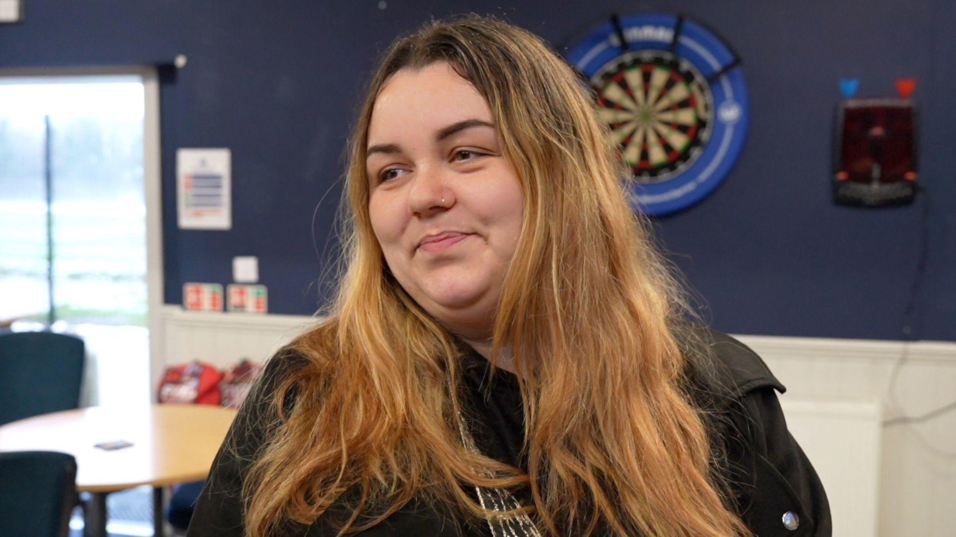 Casey Cooper pictured in a community room with a dartboard and cafe tables behind. She has a nose ring, long fair hair with darker roots, and is smiling while looking to the left of the photo.