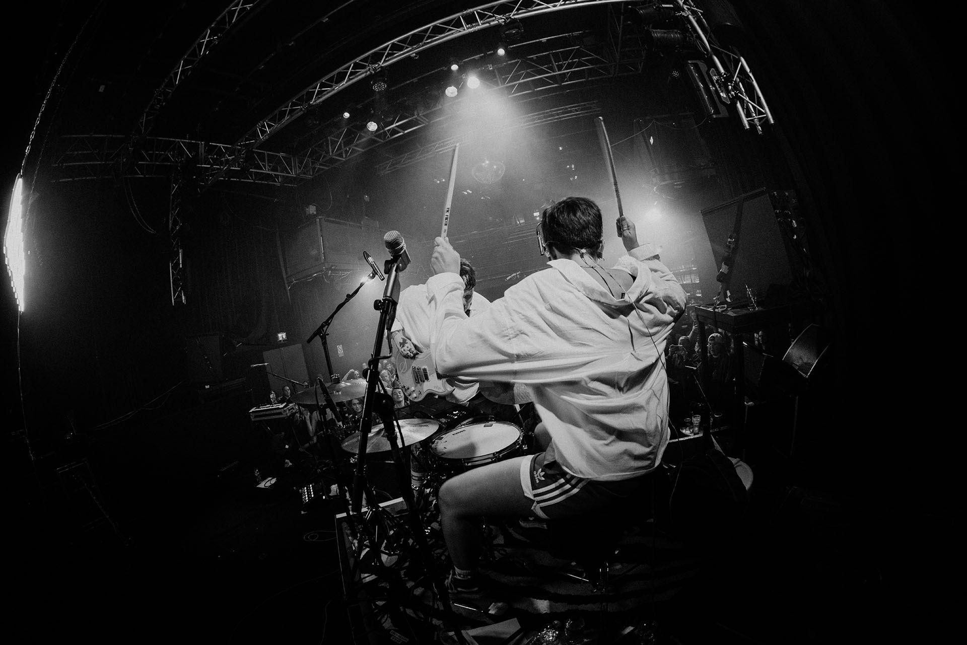 a black and white photo of arxx's drummer clara townsend. she is sat at her drum kit with her back to us, with drumsticks raised in the air. she wears a loose-fitting white t-shirt