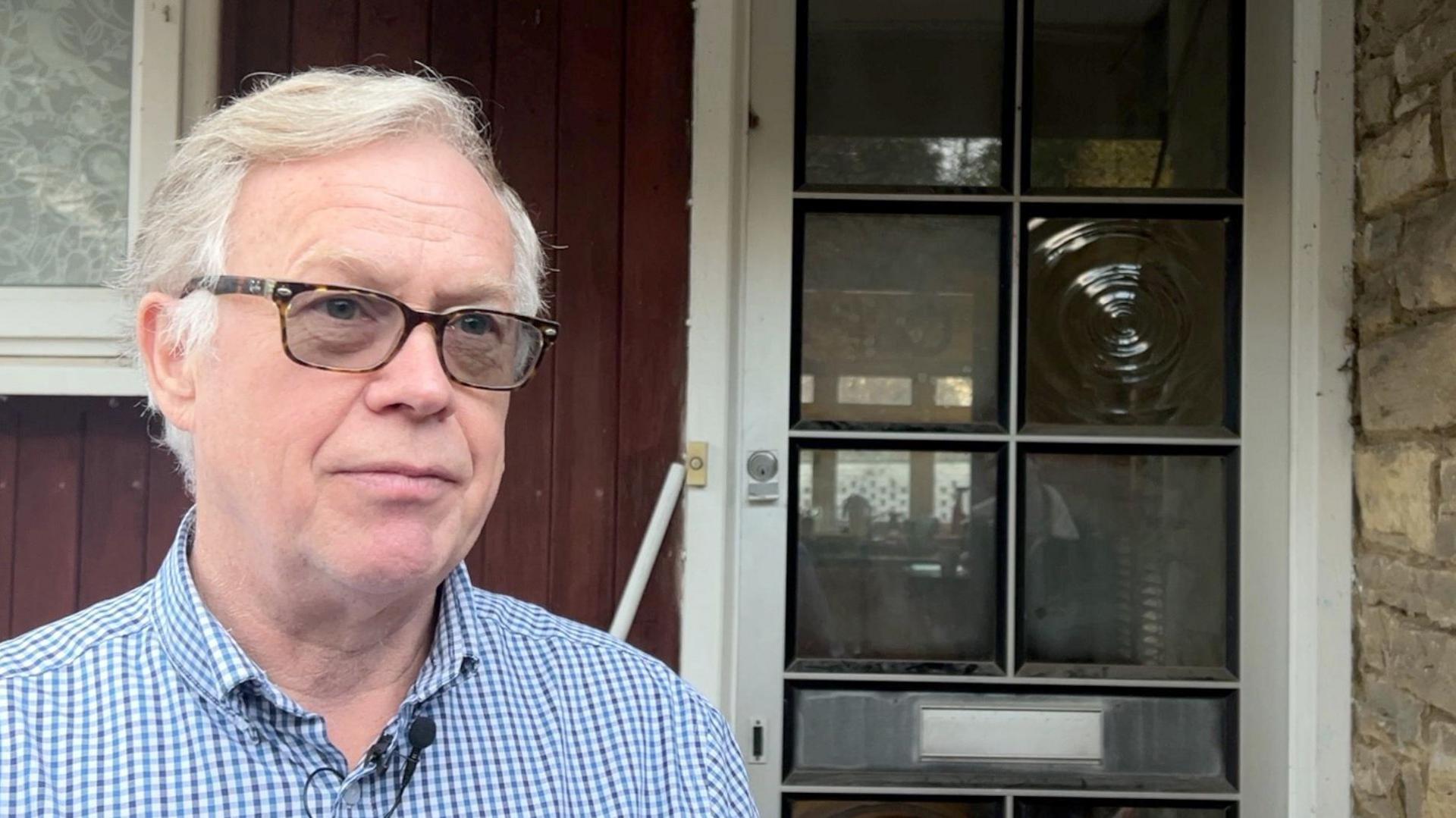 Andy Baker has short white hair and slightly darkly tinted glasses. He is wearing a blue and white checkered button shirt. He stands by the front door of his house in the left hand side of the frame, looking out of frame to the right. 