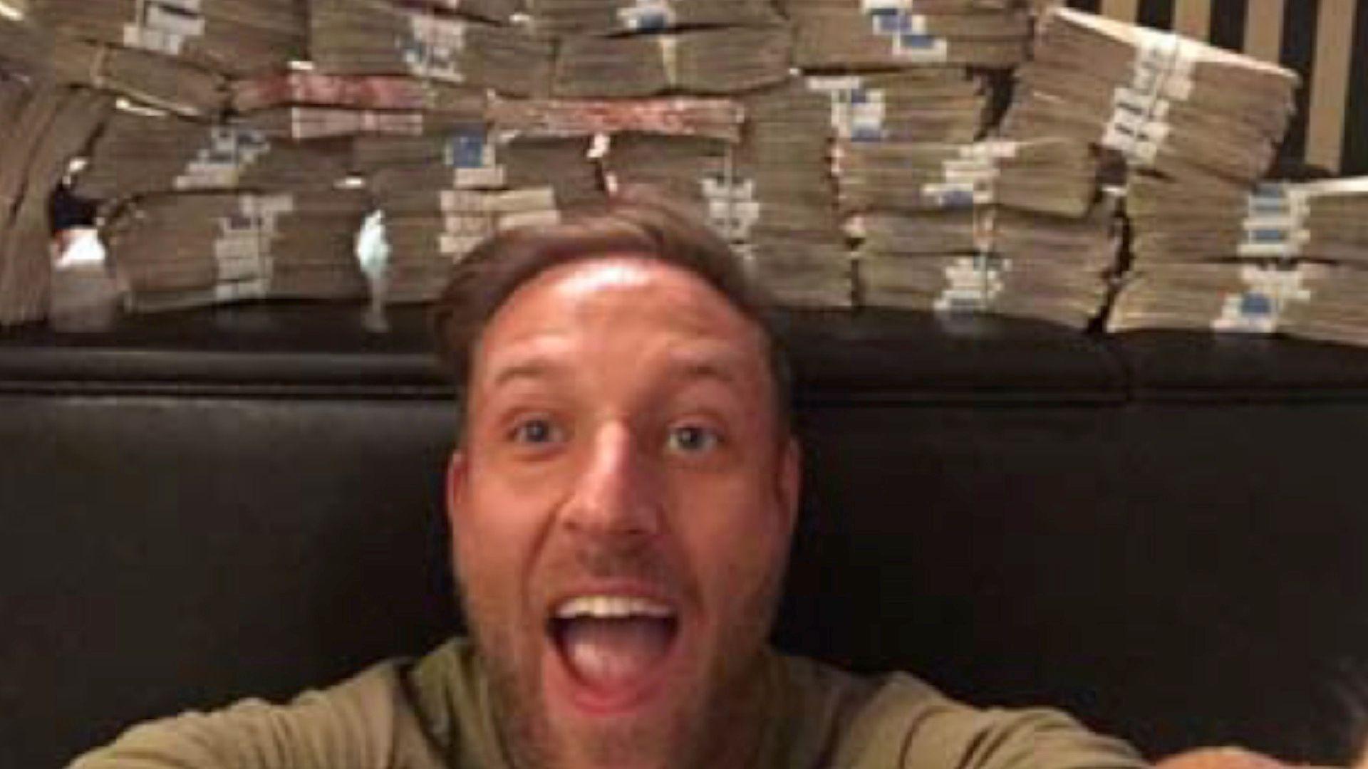 Man crouched below a large pile of money smiles for the camera.
