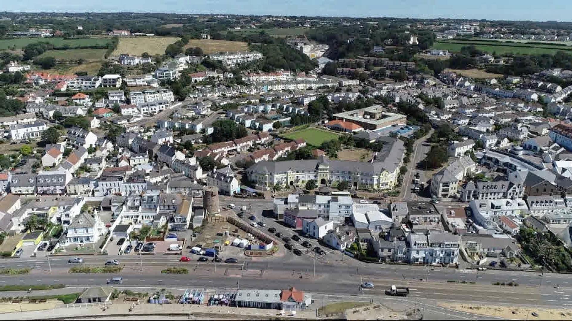 An aerial view of St Helier, with a main road in front of different blocks of buildings, fields and trees seen further afield and a line of blue sky.