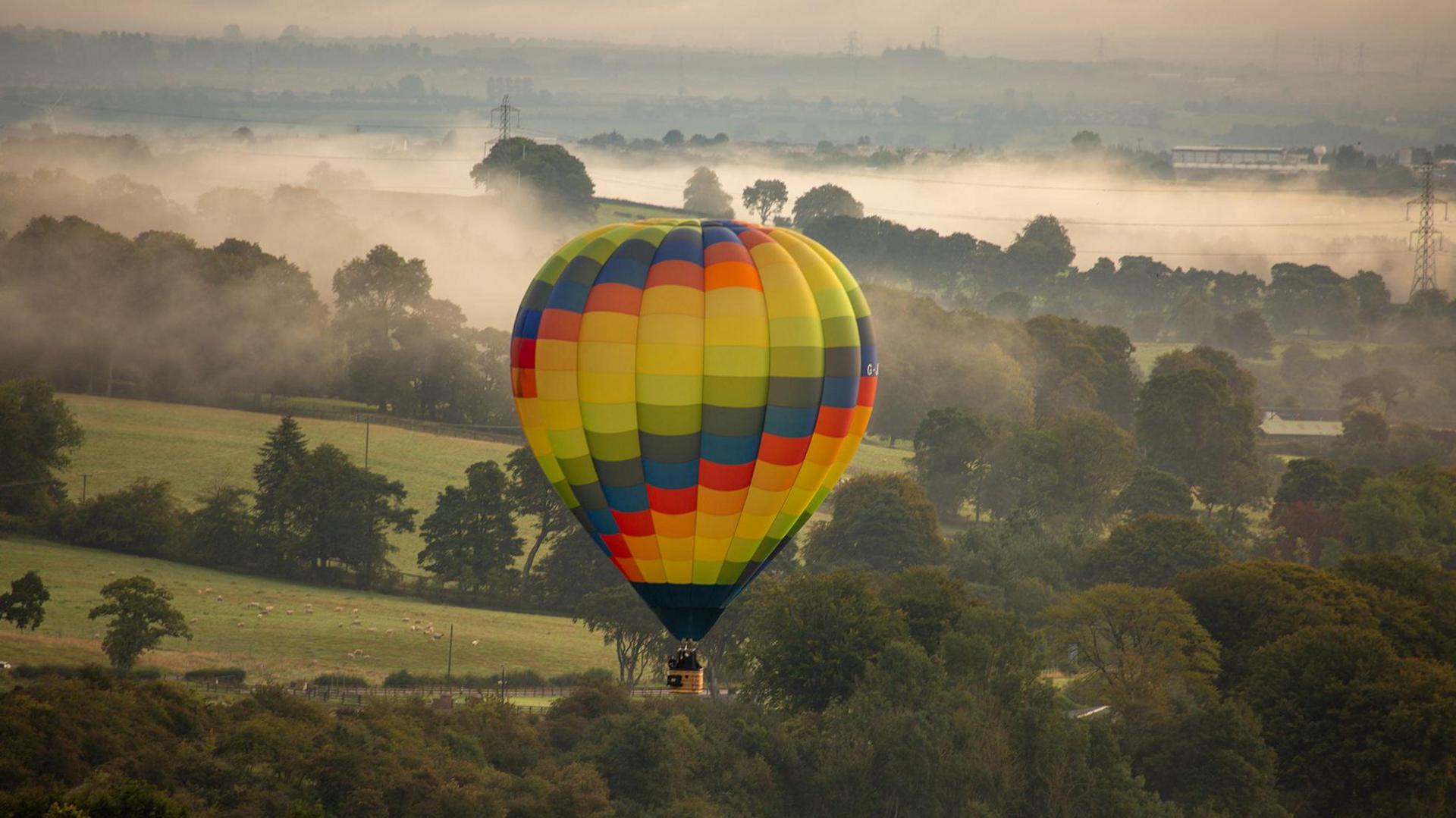 Chequered hot air balloon floats over green fields and trees