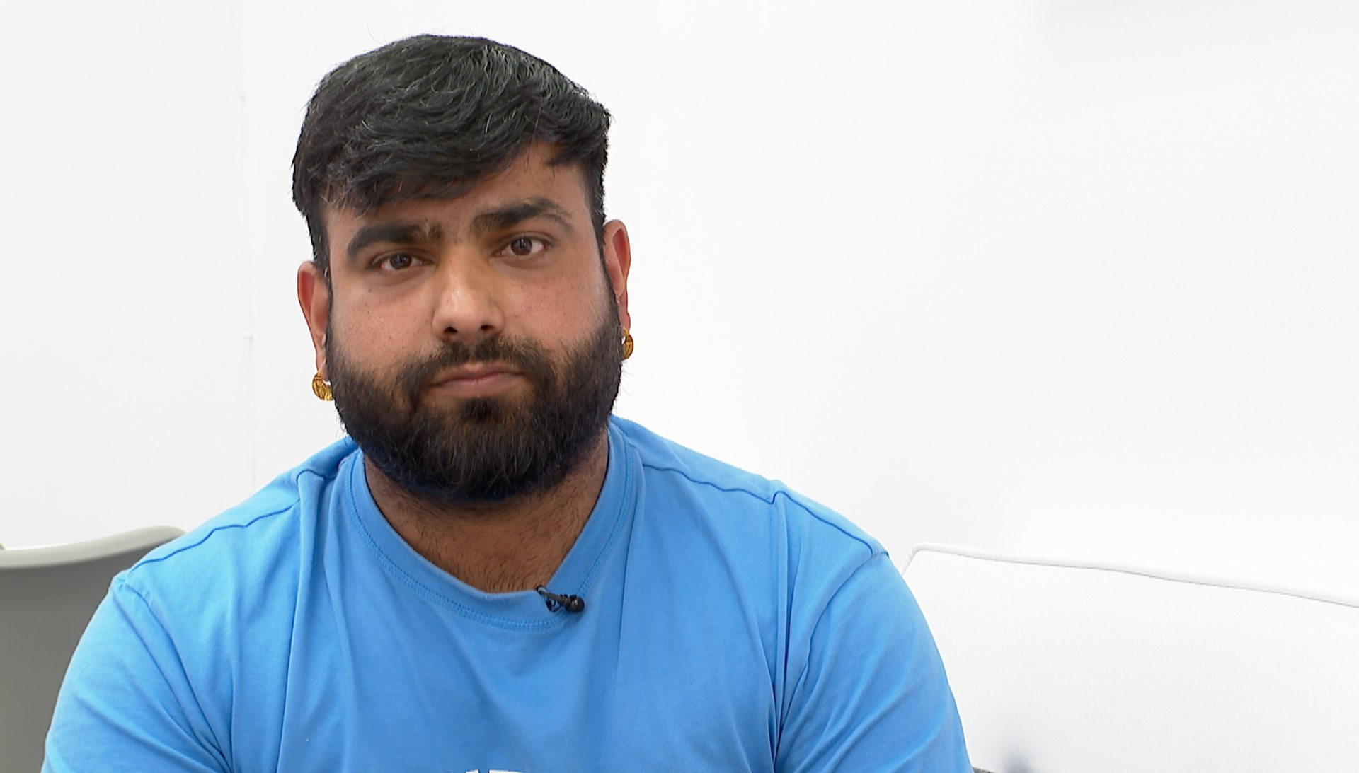 A man with short hair and a beard and moustache, wearing a blue t shirt. He is looking at the camera but is not smiling.