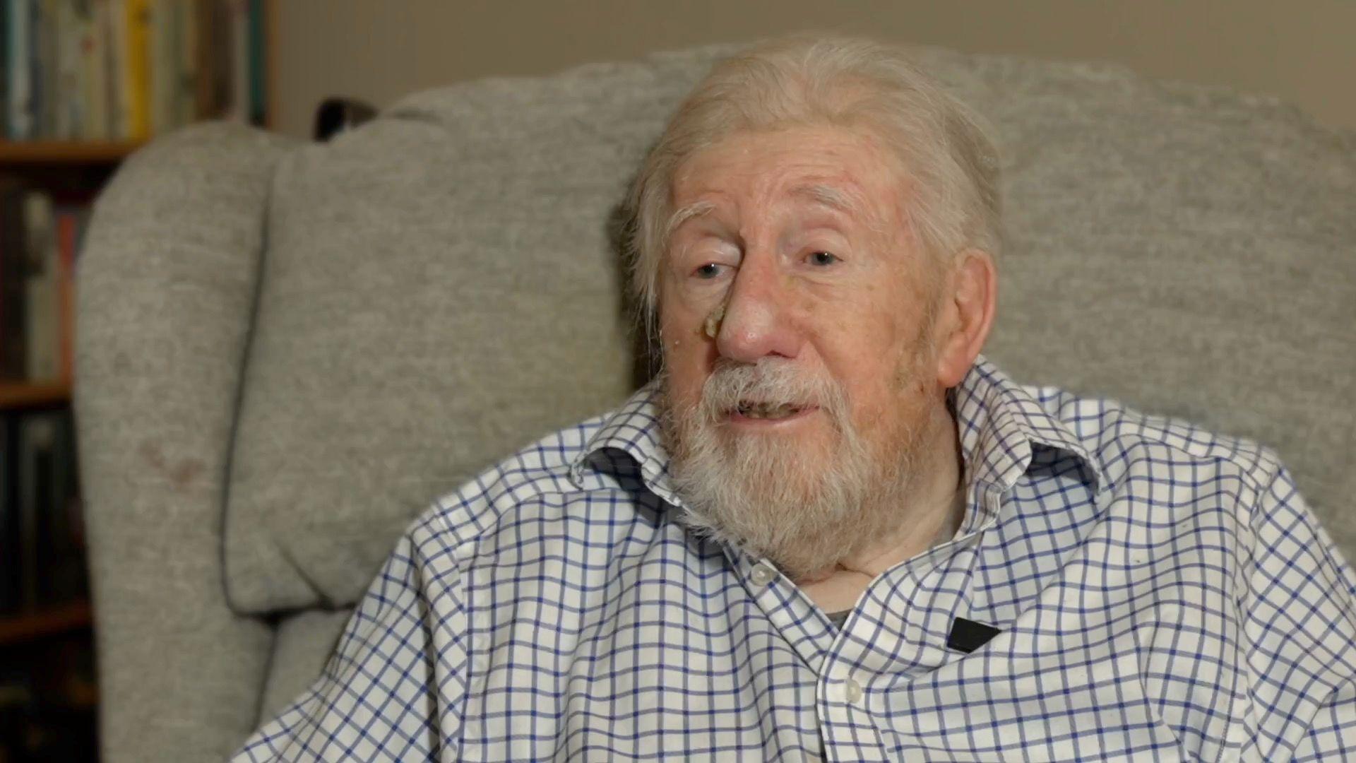 Trevor is sat on a grey sofa wearing a blue striped shirt. He is white hair and a white beard.