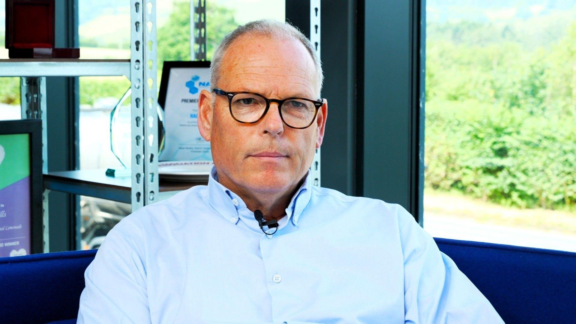William Watkin, wearing a blue shirt and glasses, has short grey hair. He has a neutral expression