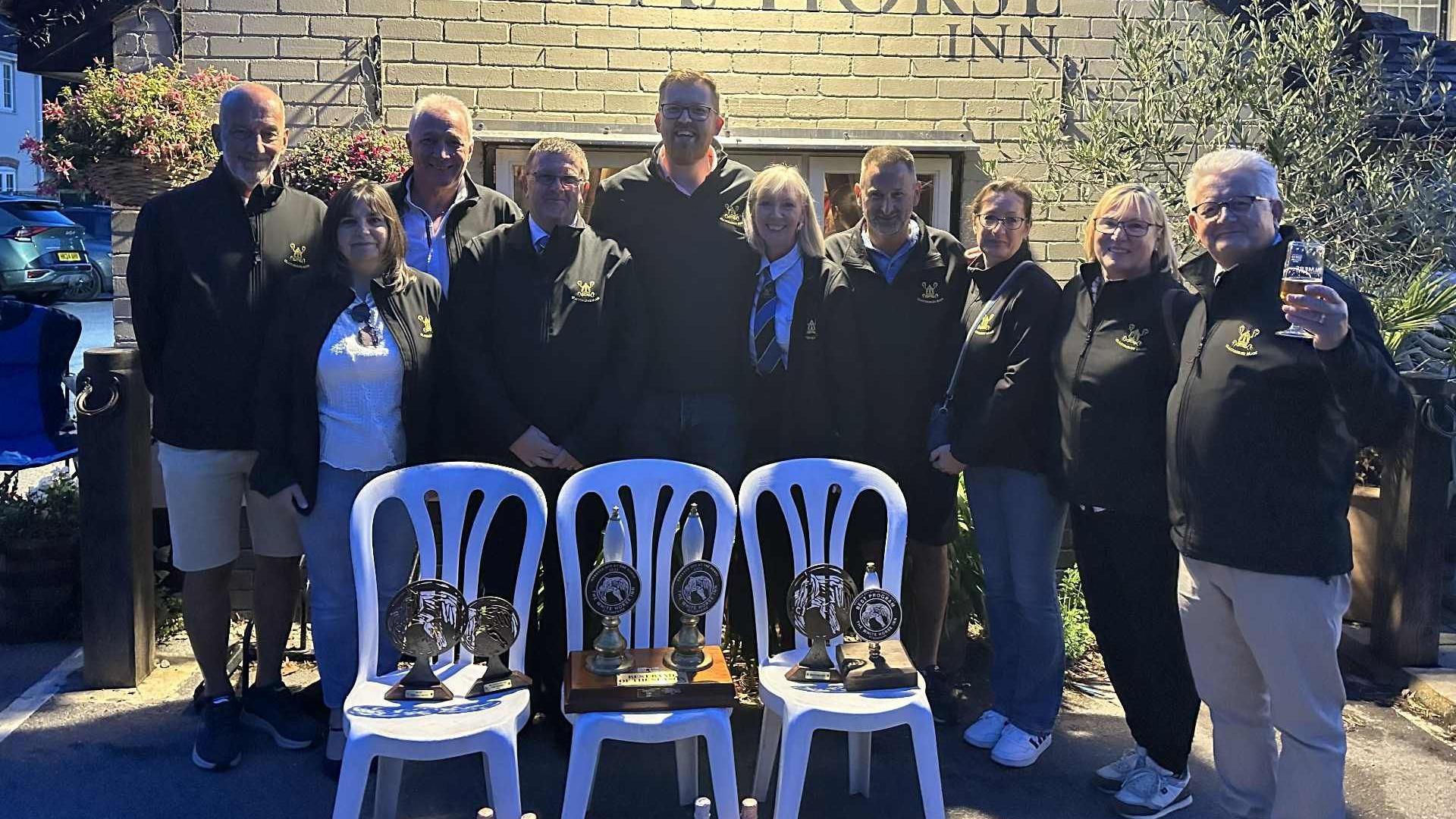 A group of people wearing black fleeces are standing next to each other smiling at the camera. They are standing behind three white plastic chairs on which they have arranged six trophies.